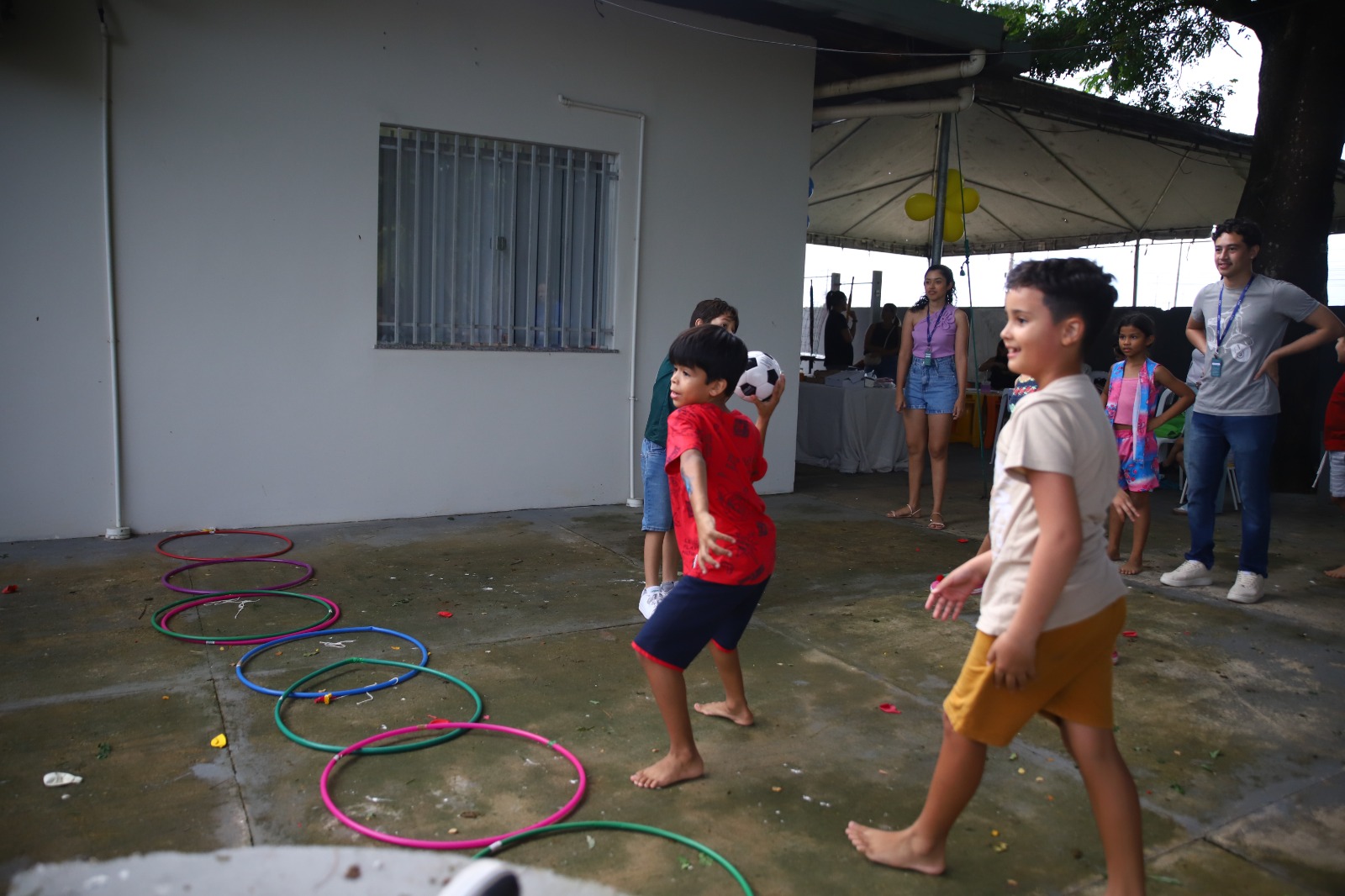 Imagem colorida mostra crianças participando de uma atividade com bola e bambolês. Um menino se prepara para arremessar a bola, enquanto outros acompanham atentos e aguardam sua vez de brincar.
