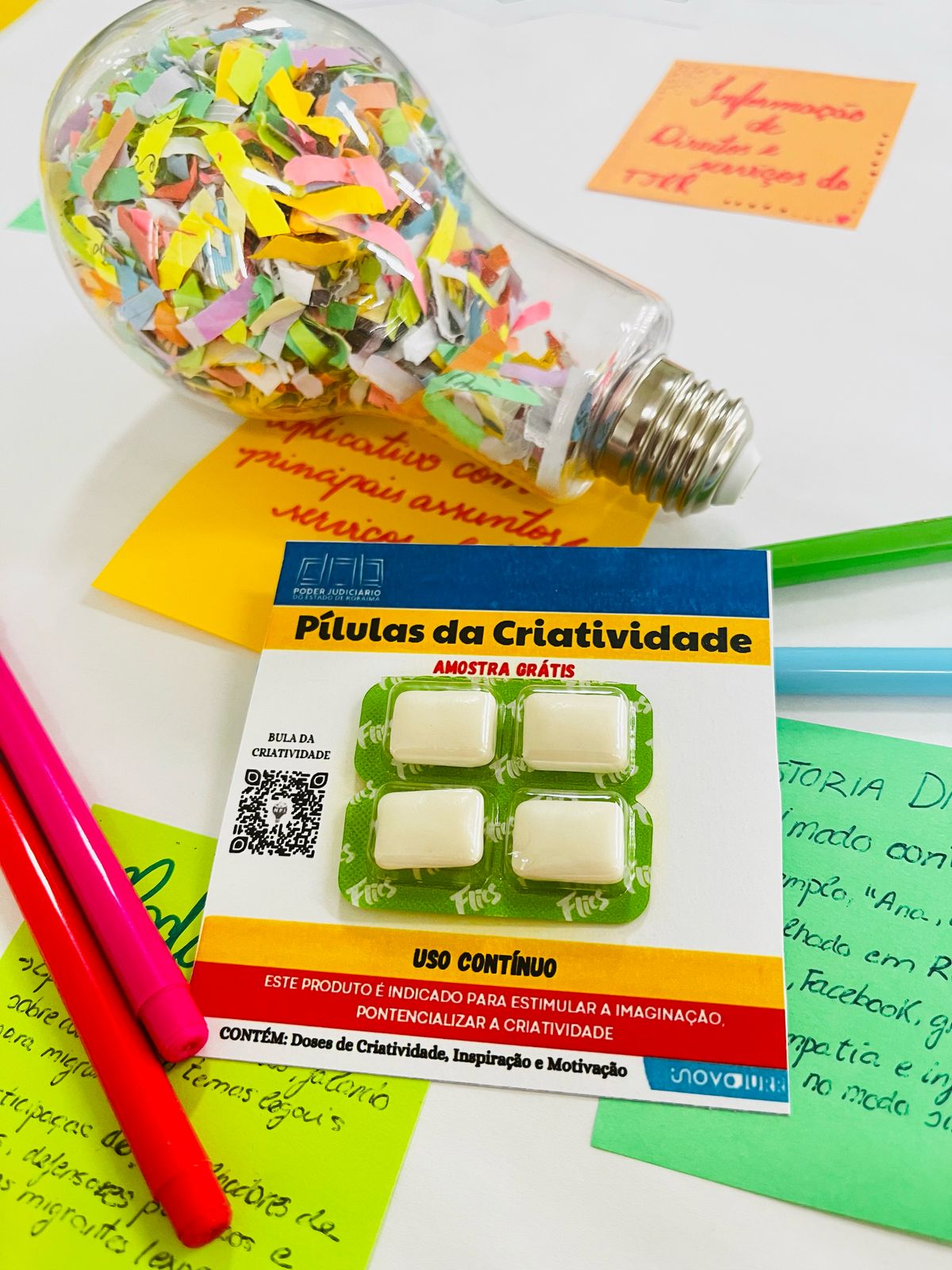  imagem colorida de uma mesa com diversos objetos relacionados à criatividade e ideias. No centro, destaca-se uma embalagem que se assemelha a um blister de remédios, intitulada "Pílulas da Criatividade". A embalagem indica ser uma "AMOSTRA GRÁTIS" e descreve o produto como sendo para "USO CONTÍNUO". Ao lado da embalagem, há uma lâmpada de vidro transparente preenchida com pequenos pedaços coloridos de papel, sugerindo a metáfora de uma "explosão" de ideias.
