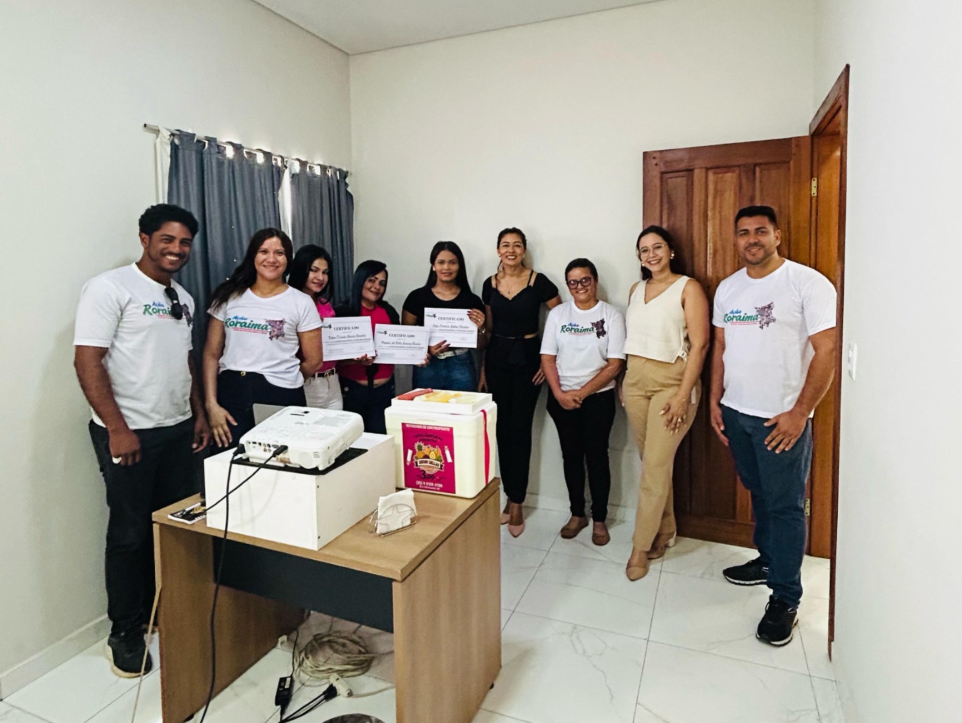  Imagem colorida mostra nove pessoas durante a capacitação do produto “Dindin Dellas”, promovida pela IARR em parceria com a CEVID/TJRR. Quatro mulheres exibem seus certificados, e à frente delas há uma mesa com um isopor contendo dindins gourmet. 