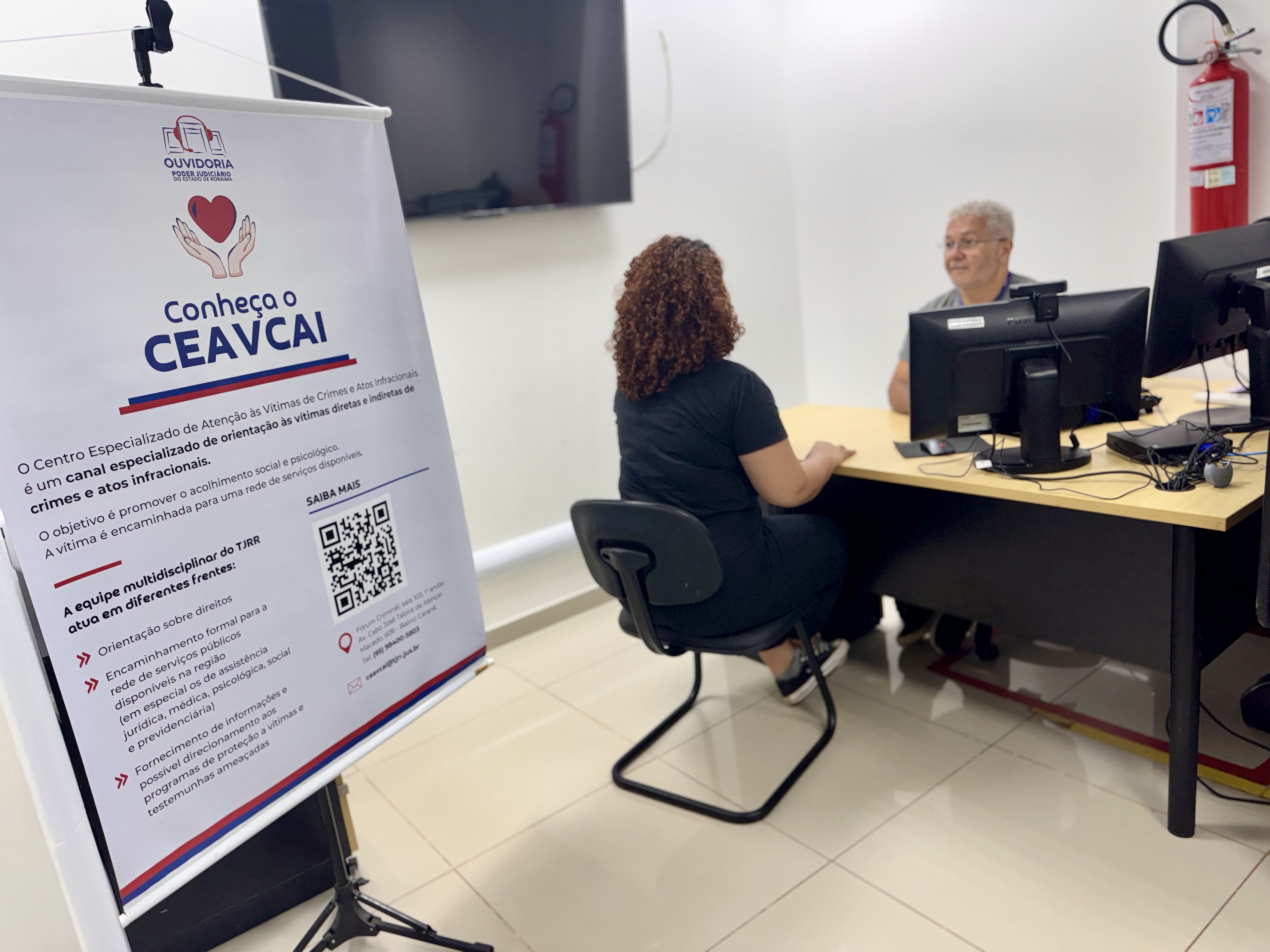 A imagem retrata um ambiente de escritório onde duas pessoas conversam diante de computadores, ao lado de um banner informativo sobre o CEAVCAI (Centro Especializado de Atendimento às Vítimas de Crimes e Atos Infracionais). O banner apresenta o serviço como um canal de orientação e acolhimento com equipe multidisciplinar, oferecendo apoio jurídico, psicológico e social, além de acesso a programas de proteção. A imagem retrata um ambiente de escritório onde duas pessoas conversam diante de computadores, ao lado de um banner informativo sobre o CEAVCAI (Centro Especializado de Atendimento às Vítimas de Crimes e Atos Infracionais). O banner apresenta o serviço como um canal de orientação e acolhimento com equipe multidisciplinar, oferecendo apoio jurídico, psicológico e social, além de acesso a programas de proteção.