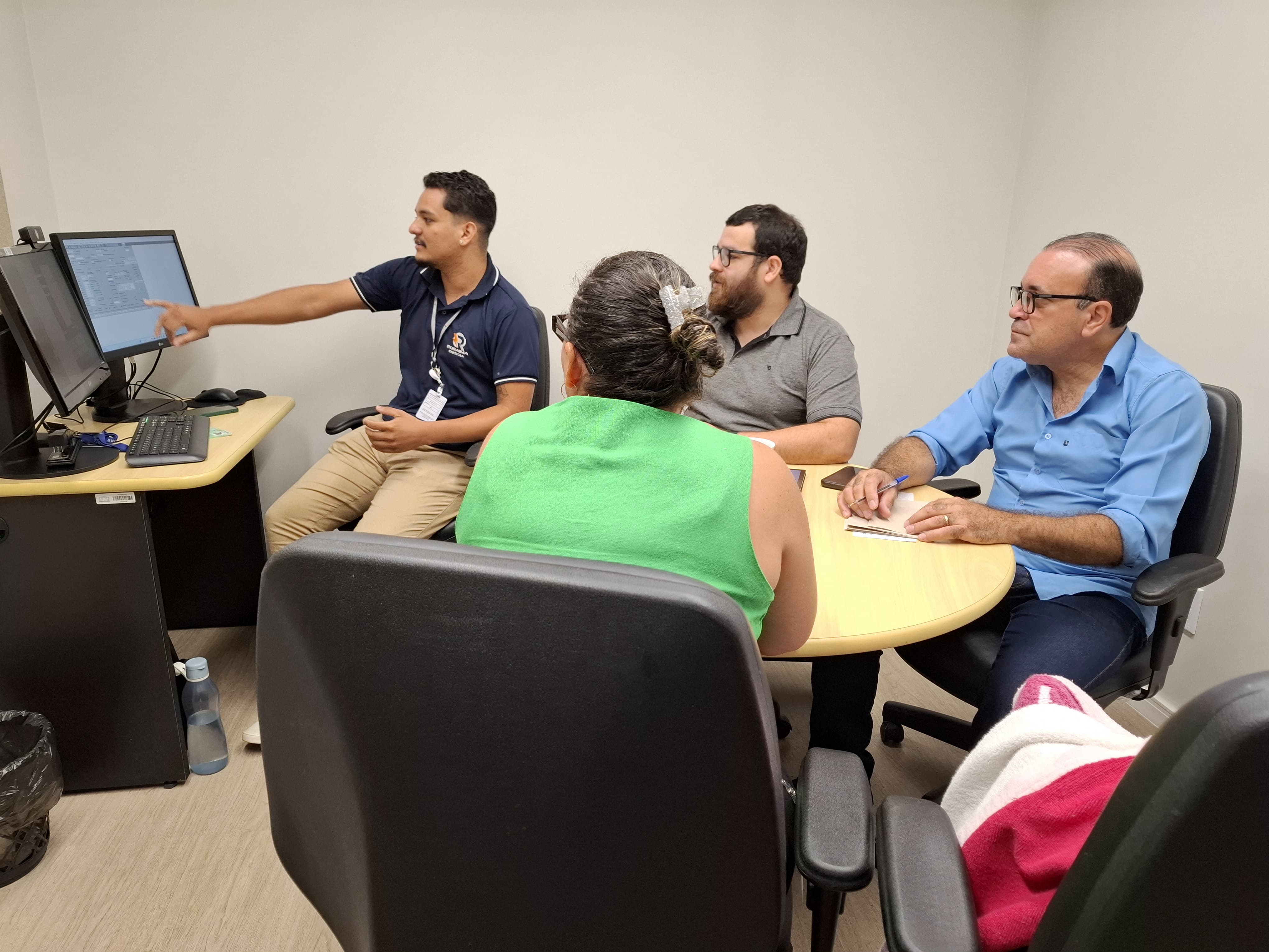 A imagem retrata um ambiente de trabalho colaborativo com quatro pessoas reunidas. Um homem aponta para a tela de um computador, enquanto os demais observam atentamente. Estão sentados em uma sala bem iluminada, sugerindo uma reunião ou atendimento técnico em equipe. A imagem retrata um ambiente de trabalho colaborativo com quatro pessoas reunidas. Um homem aponta para a tela de um computador, enquanto os demais observam atentamente. Estão sentados em uma sala bem iluminada, sugerindo uma reunião ou atendimento técnico em equipe.