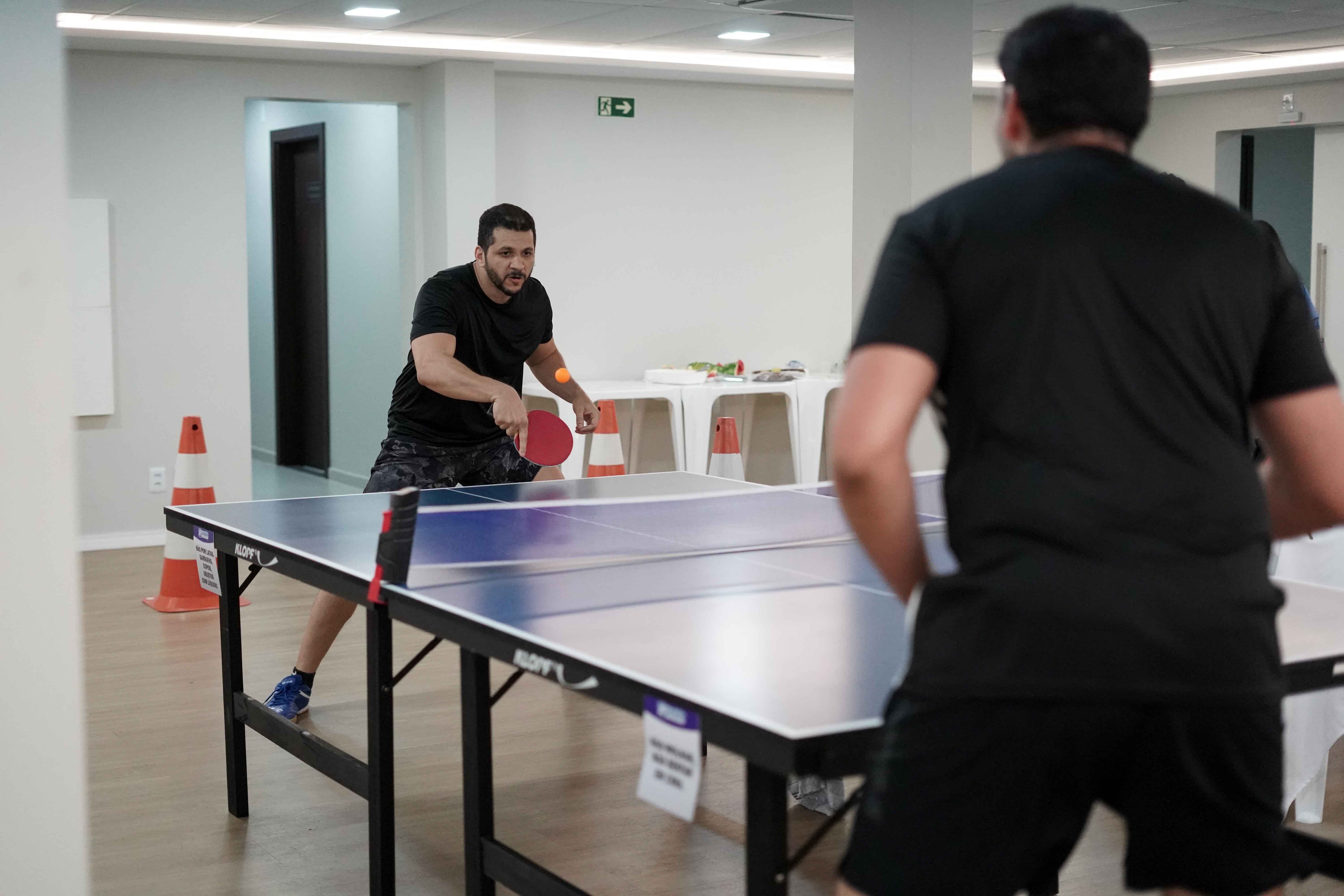A imagem mostra dois homens concentrados em uma partida de tênis de mesa em um ambiente interno e bem iluminado. Ambos usam roupas esportivas escuras, e o jogador em destaque segura uma raquete vermelha, pronto para rebater a bola laranja. A imagem mostra dois homens concentrados em uma partida de tênis de mesa em um ambiente interno e bem iluminado. Ambos usam roupas esportivas escuras, e o jogador em destaque segura uma raquete vermelha, pronto para rebater a bola laranja.
