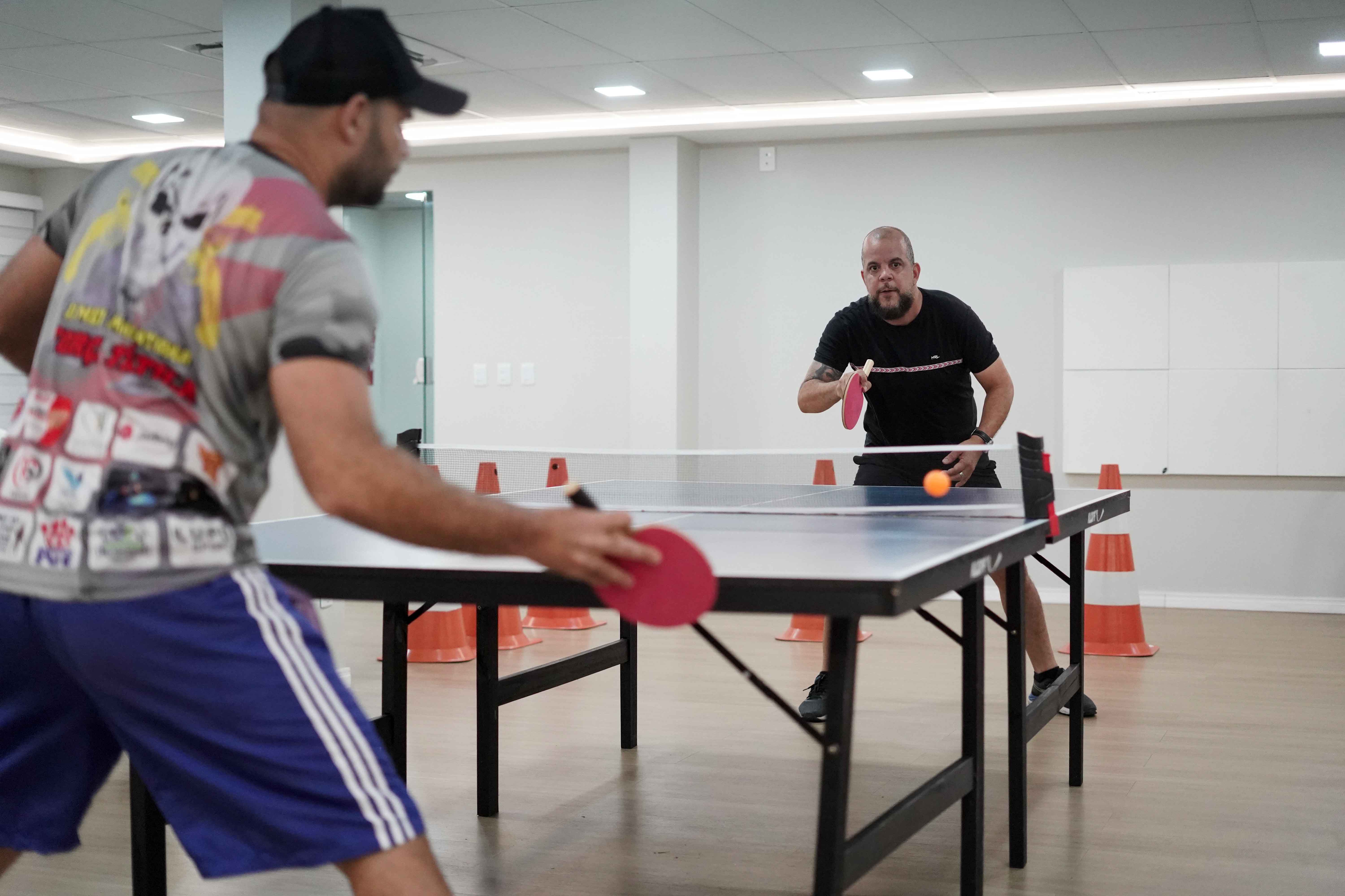 A imagem mostra uma partida de tênis de mesa entre dois homens em um ambiente interno bem iluminado. Um deles, de boné e camiseta cinza, se prepara para rebater, enquanto o adversário, de preto, concentra-se na jogada, com a bola laranja no ar. A imagem mostra uma partida de tênis de mesa entre dois homens em um ambiente interno bem iluminado. Um deles, de boné e camiseta cinza, se prepara para rebater, enquanto o adversário, de preto, concentra-se na jogada, com a bola laranja no ar.
