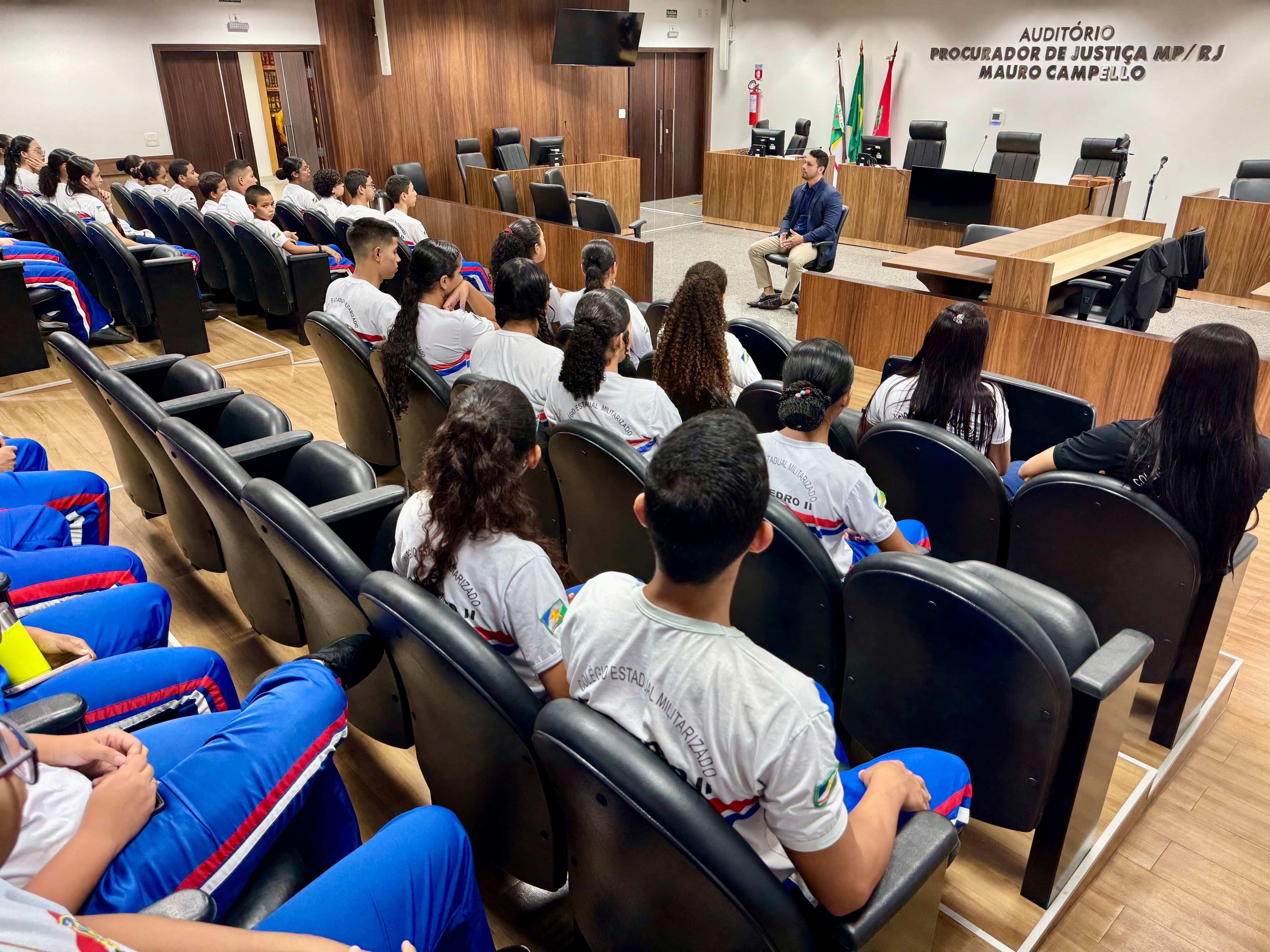 Imagem colorida mostra auditório com estudantes do Colégio Dom Pedro II, que participam de uma roda de conversa com o juiz Cleber Gonçalves. O magistrado está à frente dos alunos, sentado em uma cadeira giratória. Algumas cadeiras do auditório estão vazias.