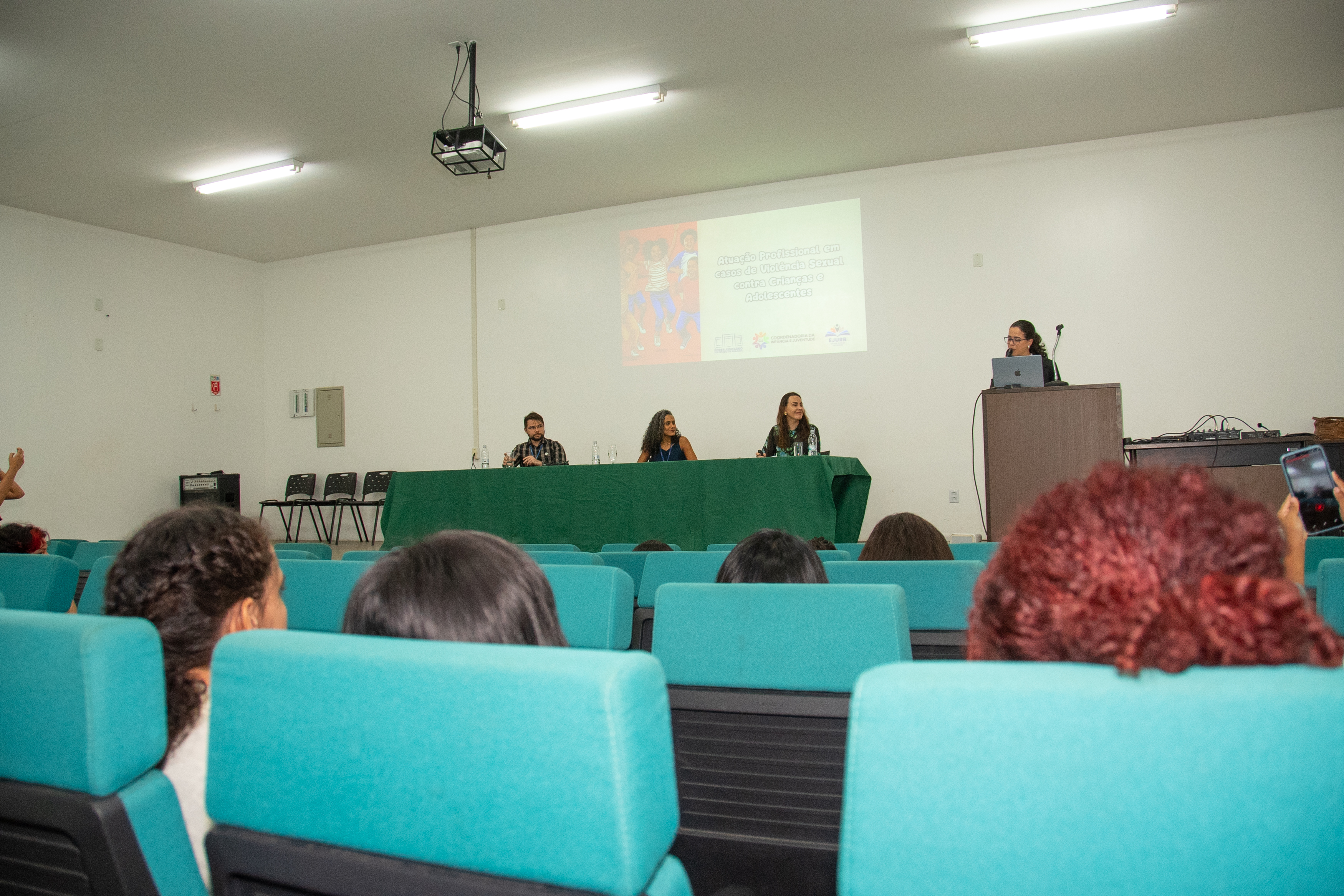 Foto colorida mostra um auditório com cadeiras verde-água ocupadas por espectadores. À frente, três pessoas estão sentadas à mesa de um painel, coberta com toalha verde, enquanto uma quarta pessoa fala em pé ao lado de um púlpito com um notebook. No fundo, é projetado um slide com o título: “Atuação Profissional em casos de Violência Sexual contra Crianças e Adolescentes”. Foto colorida mostra um auditório com cadeiras verde-água ocupadas por espectadores. À frente, três pessoas estão sentadas à mesa de um painel, coberta com toalha verde, enquanto uma quarta pessoa fala em pé ao lado de um púlpito com um notebook. No fundo, é projetado um slide com o título: “Atuação Profissional em casos de Violência Sexual contra Crianças e Adolescentes”.