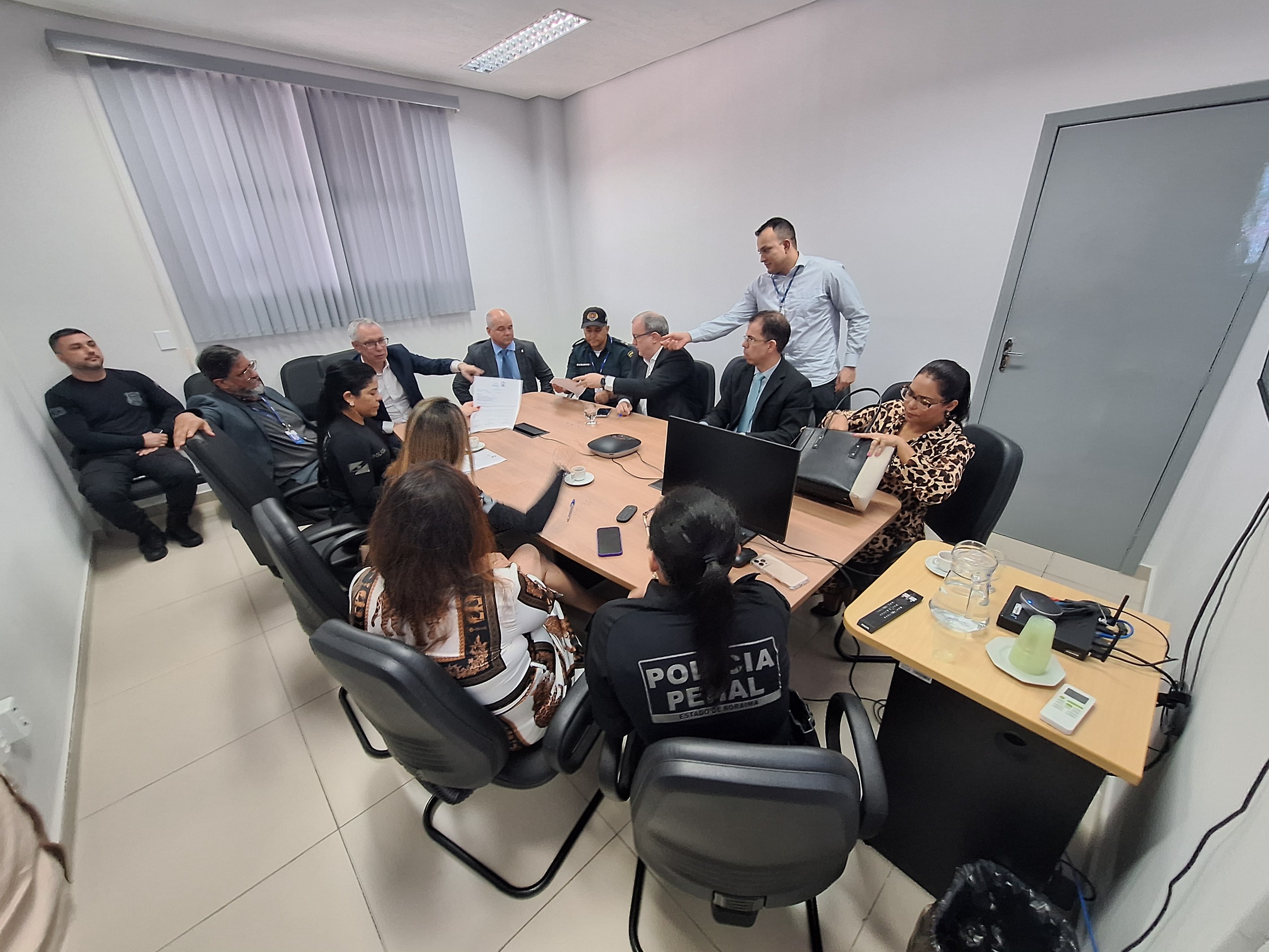 Foto colorida mostra uma reunião formal em andamento, realizada em uma sala fechada. Ao redor de uma mesa retangular estão sentadas cerca de treze pessoas, entre elas homens e mulheres, com diferentes trajes — alguns em ternos, outros com uniformes da Polícia Penal. Foto colorida mostra uma reunião formal em andamento, realizada em uma sala fechada. Ao redor de uma mesa retangular estão sentadas cerca de treze pessoas, entre elas homens e mulheres, com diferentes trajes — alguns em ternos, outros com uniformes da Polícia Penal.