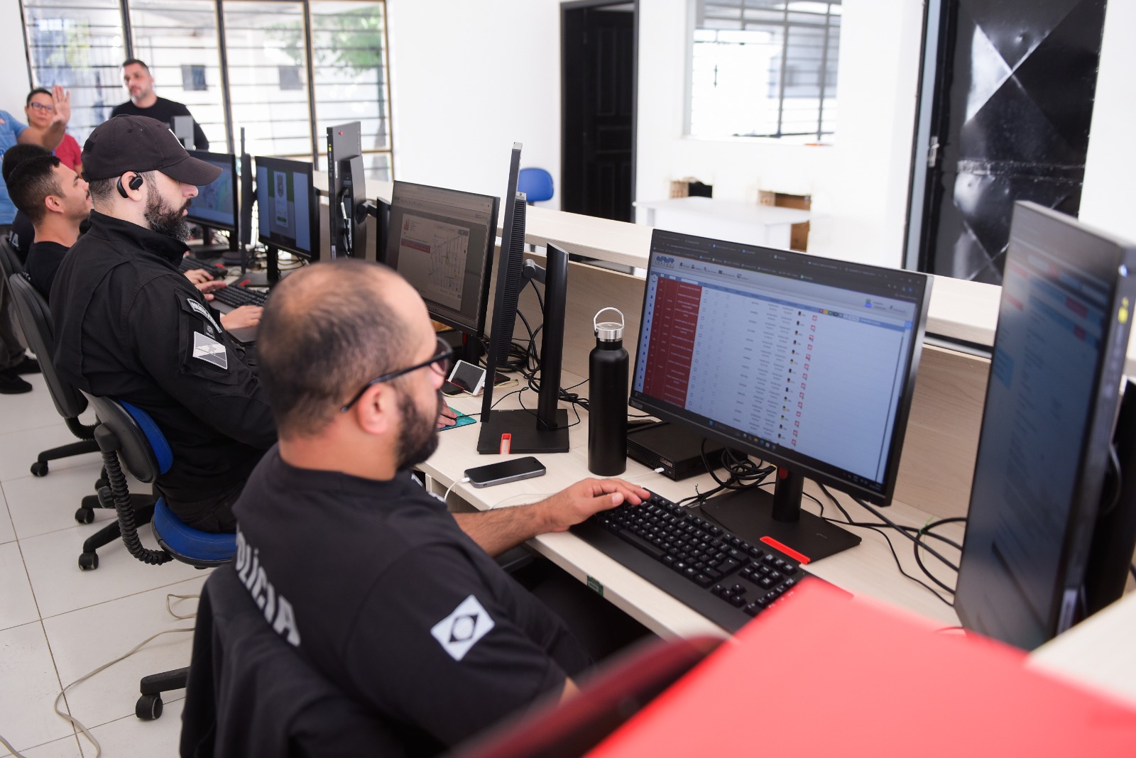 Imagem colorida mostra dois homens sentados em cadeiras de escritório em frente a computadores. Imagem colorida mostra dois homens sentados em cadeiras de escritório em frente a computadores.