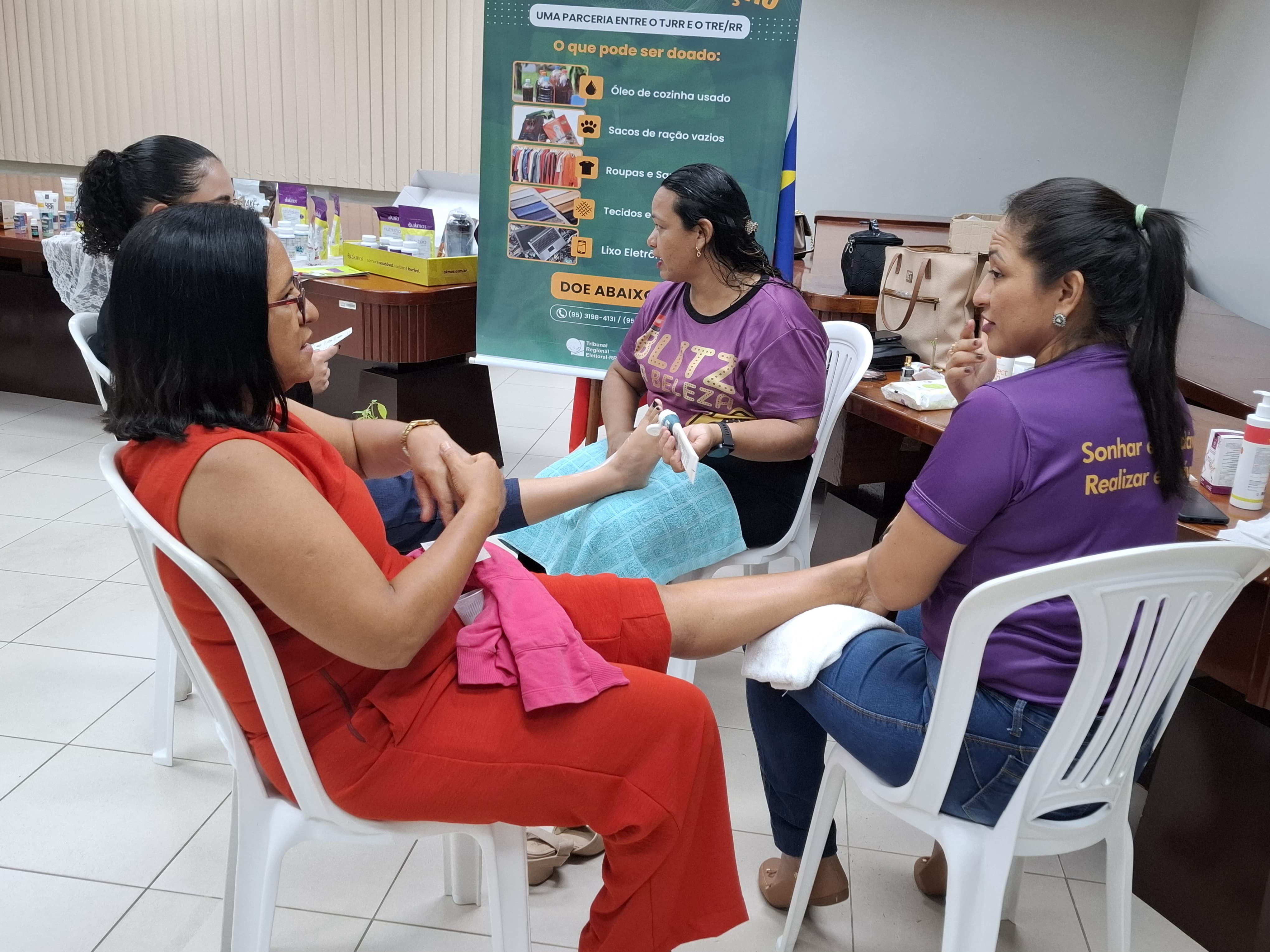 Imagem colorida mostra quatro mulheres em uma sala. Duas mulheres estão sentadas em cadeiras brancas, uma de vestido vermelho e outra de camiseta roxa, recebendo algum tipo de tratamento nos pés de outras duas mulheres que estão ajoelhadas ou curvadas em frente a elas. Ao fundo, há um banner verde com informações sobre "Uma parceria entre o TJRR e o TRE/RR" e uma lista de itens que podem ser doados, como "óleo de cozinha usado", "sacos de ração vazios", "roupas e sapatos", "tecidos e retalhos" e "lixo eletrônico".