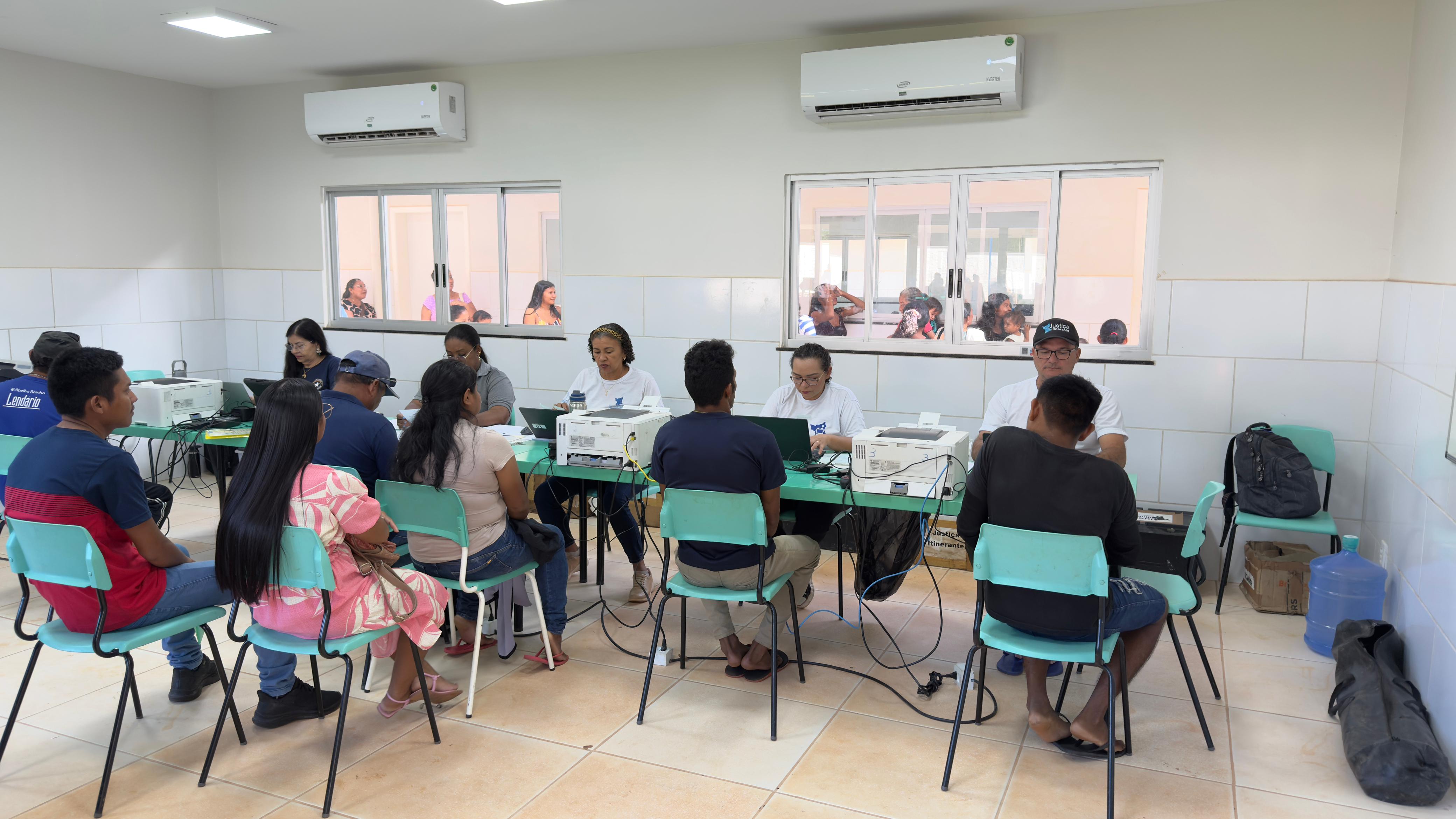 A imagem colorida mostra um ambiente interno, que parece ser uma sala de aula ou um escritório, com várias pessoas sentadas em cadeiras voltadas para mesas. Em cada mesa, há pelo menos um laptop e uma impressora, sugerindo um local de trabalho ou atendimento. As pessoas sentadas à frente parecem estar auxiliando ou interagindo com as pessoas que estão sentadas de costas para a câmera. Há janelas na parede do fundo, através das quais é possível ver mais pessoas e, possivelmente, um ambiente externo ou outra sala. Aparelhos de ar condicionado estão instalados acima das janelas. A imagem colorida mostra um ambiente interno, que parece ser uma sala de aula ou um escritório, com várias pessoas sentadas em cadeiras voltadas para mesas. Em cada mesa, há pelo menos um laptop e uma impressora, sugerindo um local de trabalho ou atendimento. As pessoas sentadas à frente parecem estar auxiliando ou interagindo com as pessoas que estão sentadas de costas para a câmera. Há janelas na parede do fundo, através das quais é possível ver mais pessoas e, possivelmente, um ambiente externo ou outra sala. Aparelhos de ar condicionado estão instalados acima das janelas.