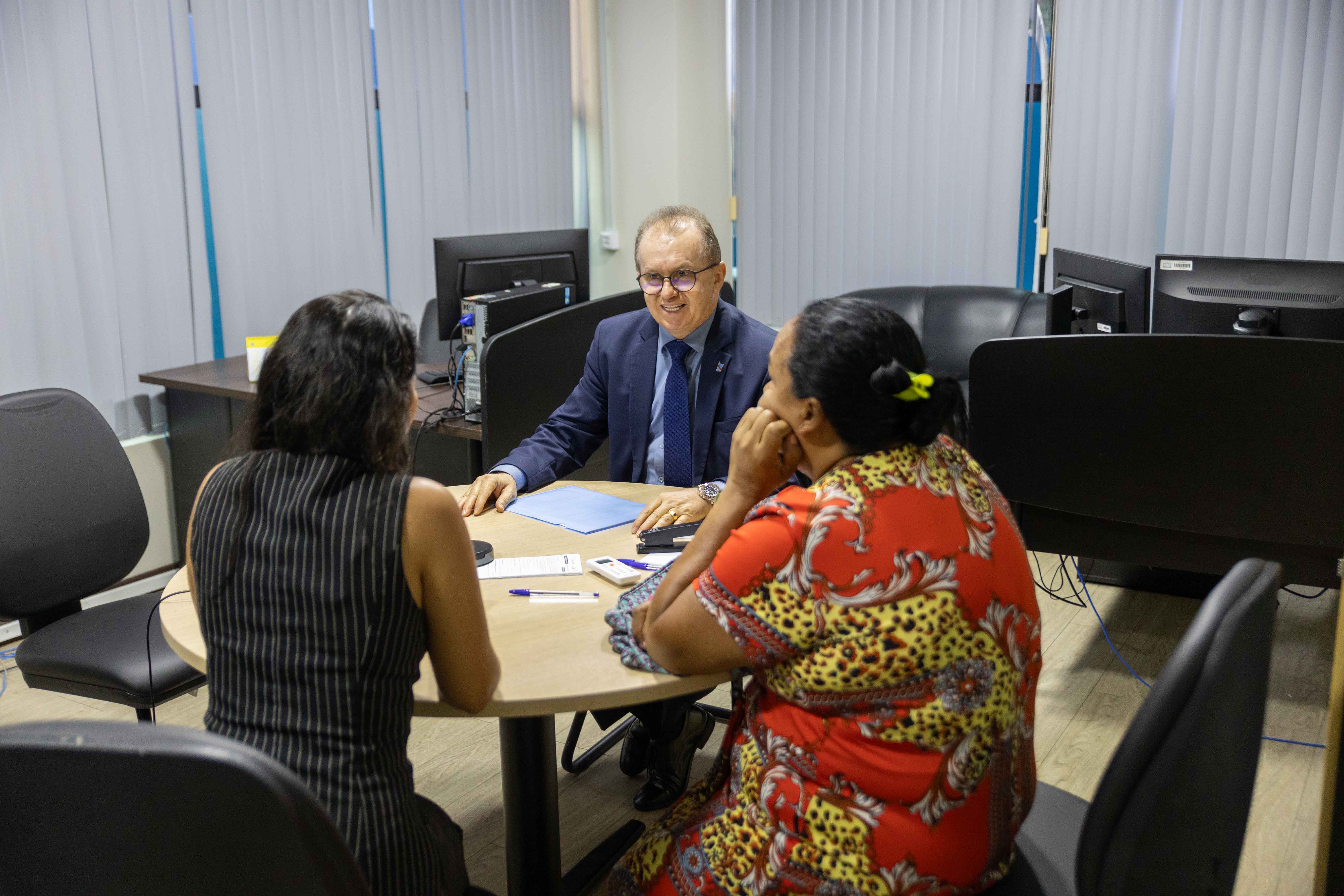 Foto colorida mostra três pessoas sentadas a mesa conversando. Um homem vestido com terno azul escuro e gravata, conversa com duas mulheres em uma sala com ambiente formal. Todos estão sentados em torno de uma mesa redonda, participando de uma audiência de conciliação. Foto colorida mostra três pessoas sentadas a mesa conversando. Um homem vestido com terno azul escuro e gravata, conversa com duas mulheres em uma sala com ambiente formal. Todos estão sentados em torno de uma mesa redonda, participando de uma audiência de conciliação.