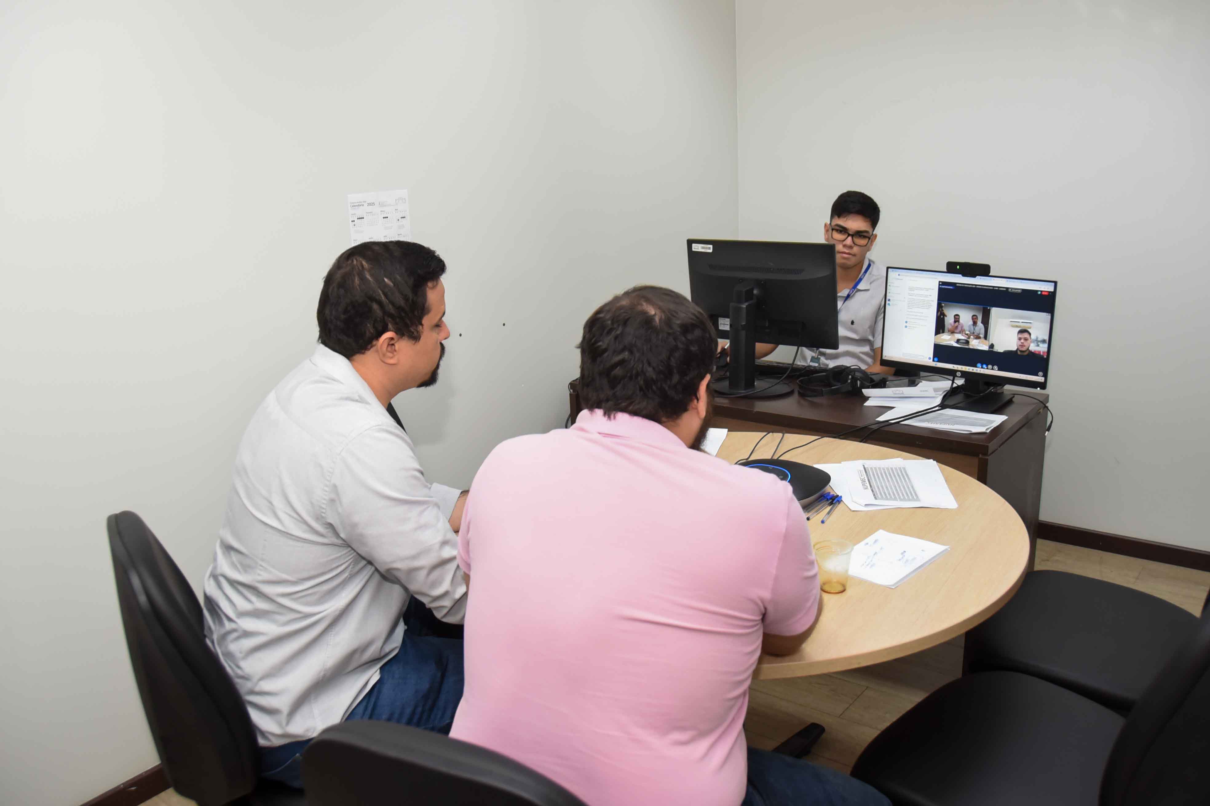 Foto colorida mostra três homens participando de uma audiência de mediação híbrida. Dois estão sentados presencialmente em frente a um servidor que conduz a sessão, enquanto outras pessoas aparecem em uma chamada de vídeo na tela do computador. Foto colorida mostra três homens participando de uma audiência de mediação híbrida. Dois estão sentados presencialmente em frente a um servidor que conduz a sessão, enquanto outras pessoas aparecem em uma chamada de vídeo na tela do computador.