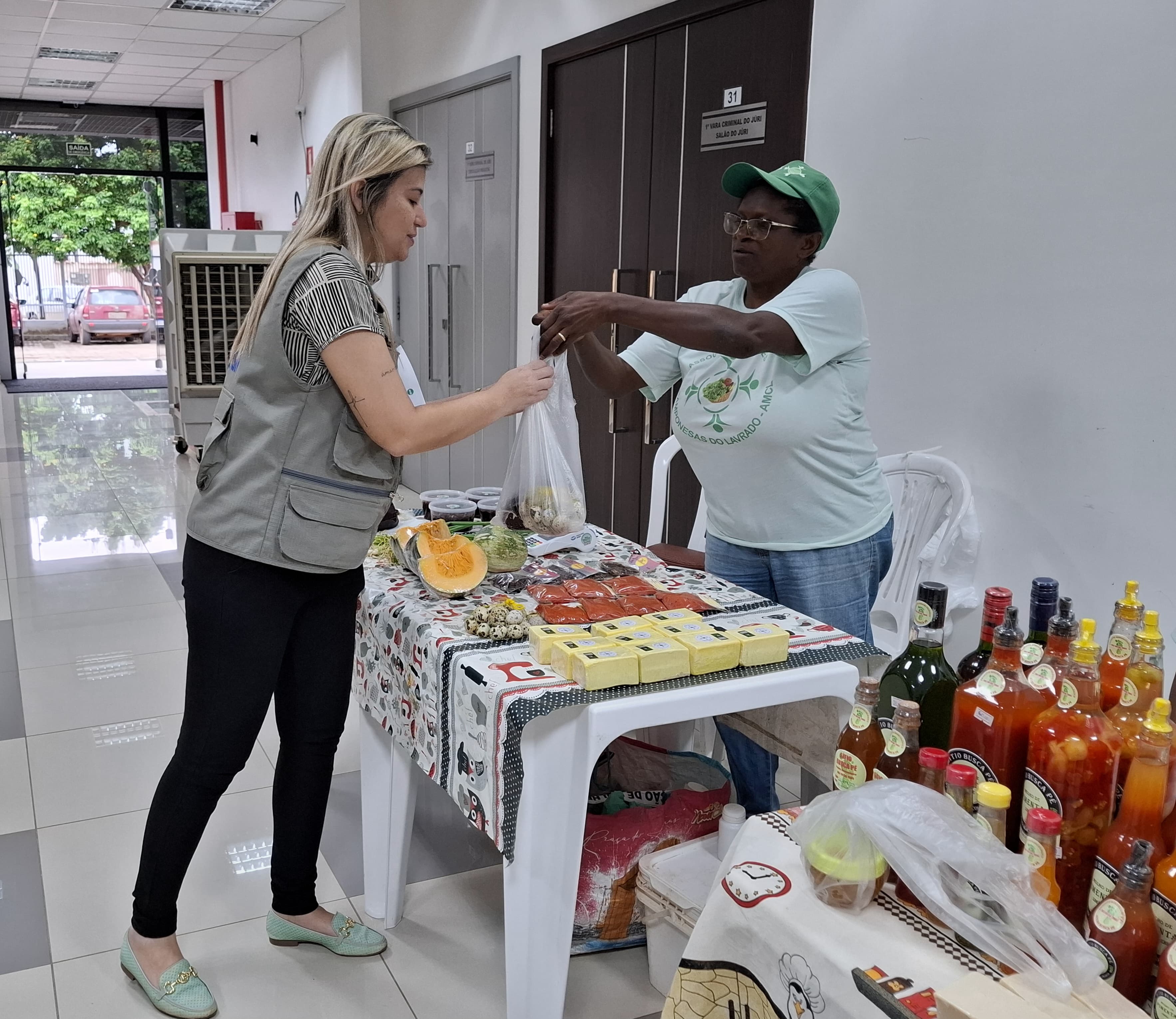 A imagem colorida mostra uma mulher entregando uma sacola com produtos para uma outra mulher de colete. Ambas estão ao redor de uma mesa, sobre a qual há diversos produtos, incluindo queijos embalados e garrafas de bebidas.