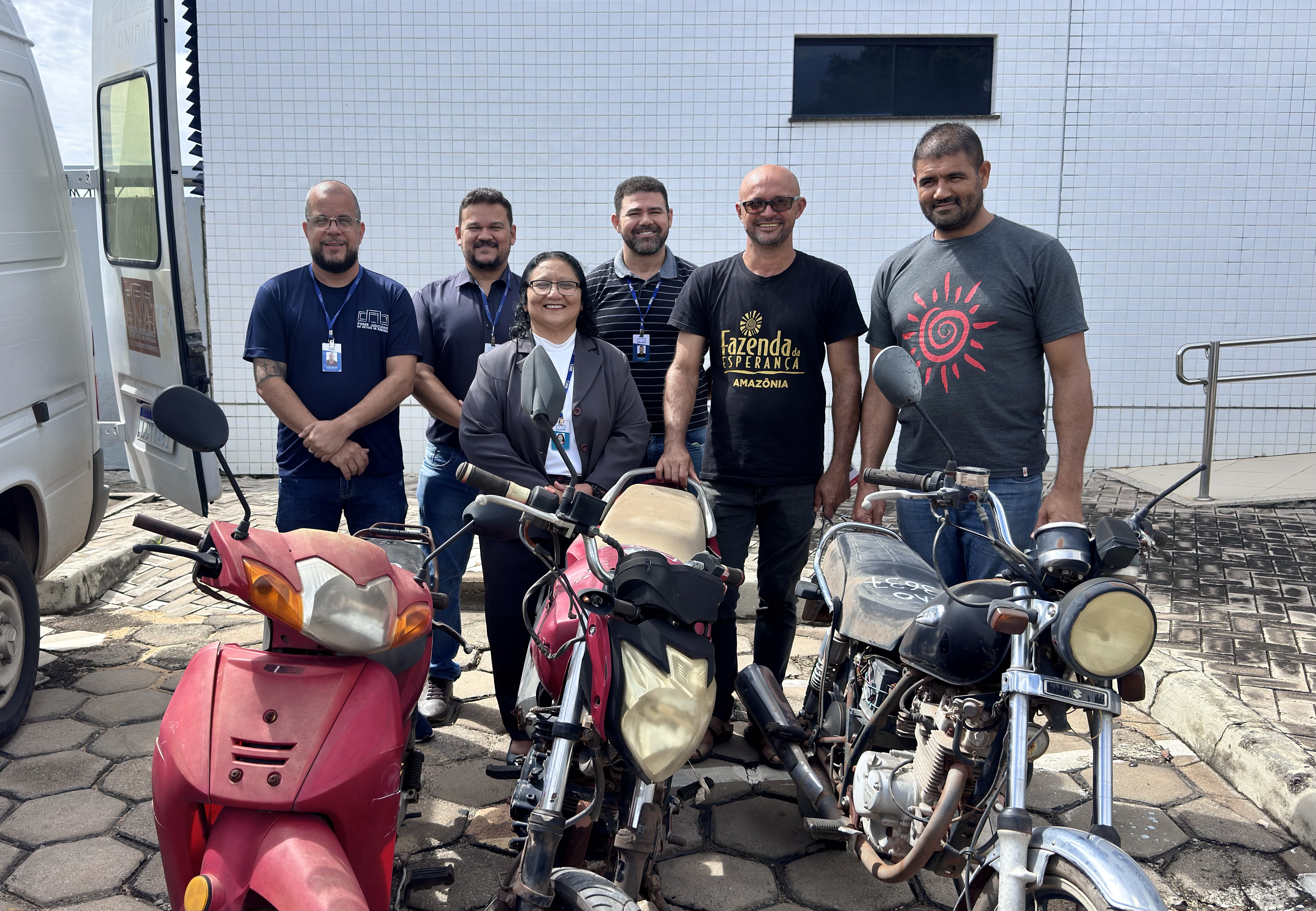Imagem colorida mostra seis pessoas estão em pé, sorrindo, posicionadas atrás de três motocicletas antigas. À esquerda, há uma van branca. Ao fundo, uma parede de azulejos brancos com uma pequena janela escura. Duas das pessoas vestem camisetas com a inscrição “Fazenda da Esperança Amazônia”, e os demais usam crachás de identificação. A cena acontece ao ar livre, em um pátio com piso de pedras.