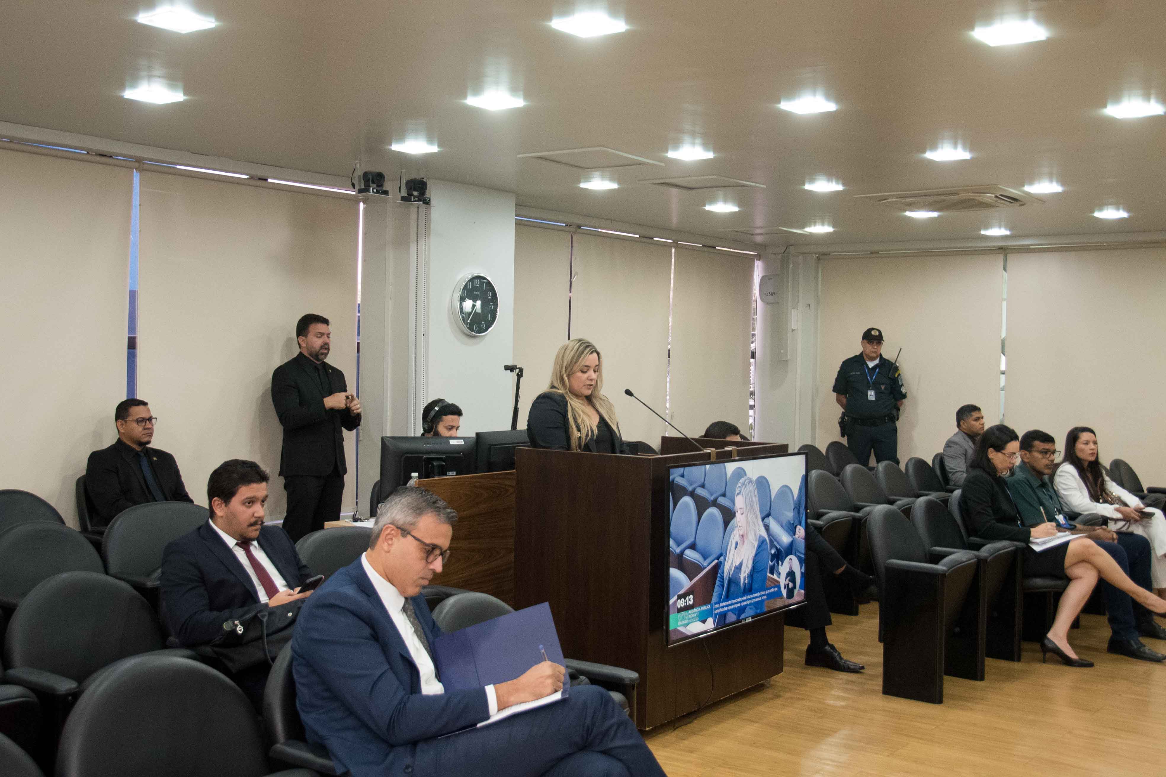 Foto de um auditório ou sala de plenário. Ao centro, uma mulher loira está de pé em uma tribuna de madeira, discursando. À sua frente, uma plateia de homens e mulheres com roupas formais assiste, sentada em cadeiras escuras. Em primeiro plano, um homem de óculos e terno anota em uma pasta. Foto de um auditório ou sala de plenário. Ao centro, uma mulher loira está de pé em uma tribuna de madeira, discursando. À sua frente, uma plateia de homens e mulheres com roupas formais assiste, sentada em cadeiras escuras. Em primeiro plano, um homem de óculos e terno anota em uma pasta.
