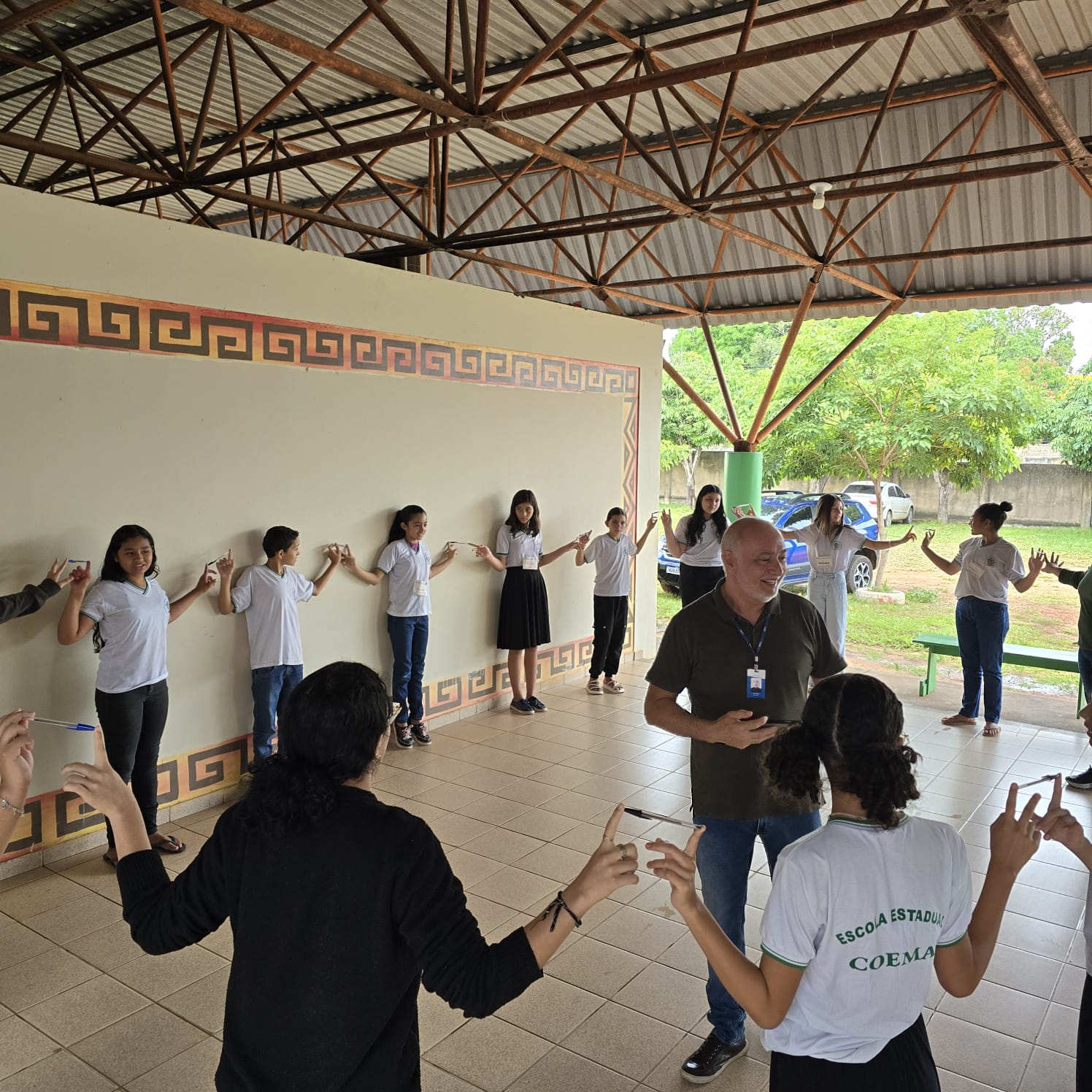 A imagem colorida mostra um grupo de cerca de 15 pessoas, incluindo adultos e jovens, participando de uma atividade em um espaço coberto. A maioria dos jovens veste uniformes escolares, e alguns adultos parecem ser educadores. As pessoas estão em círculo, algumas com os braços estendidos e segurando canetas, sugerindo um jogo ou exercício de equipe. Há uma parede clara com um padrão geométrico decorativo e, ao fundo, árvores e um carro. A imagem colorida mostra um grupo de cerca de 15 pessoas, incluindo adultos e jovens, participando de uma atividade em um espaço coberto. A maioria dos jovens veste uniformes escolares, e alguns adultos parecem ser educadores. As pessoas estão em círculo, algumas com os braços estendidos e segurando canetas, sugerindo um jogo ou exercício de equipe. Há uma parede clara com um padrão geométrico decorativo e, ao fundo, árvores e um carro.