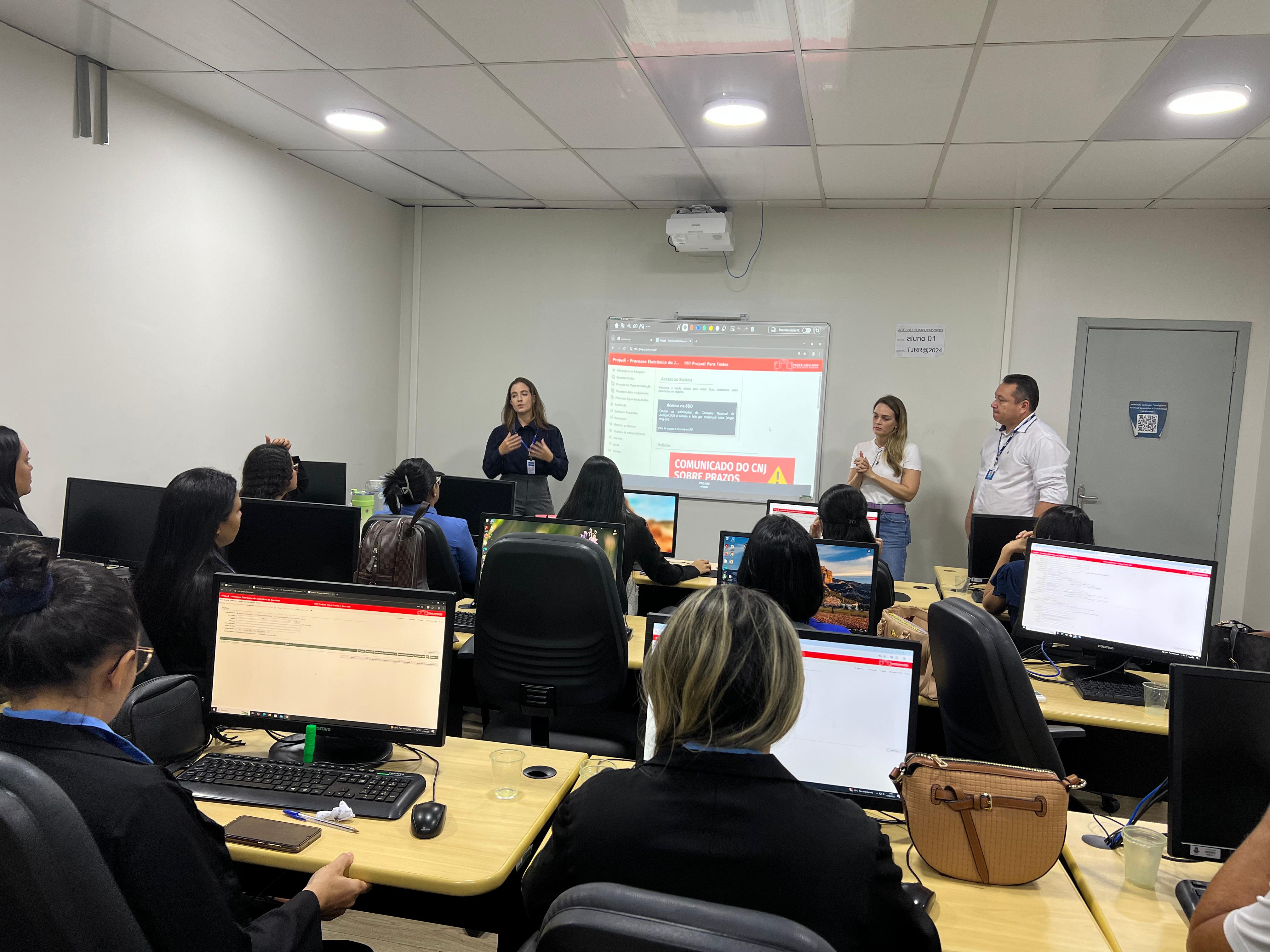 Foto colorida mostra várias pessoas em uma sala de informática participando de uma capacitação. Três instrutores estão em pé à frente da sala — duas mulheres e um homem — enquanto uma das facilitadoras fala aos participantes. No ambiente, todos os alunos estão sentados diante de computadores, acompanhando o conteúdo projetado na parede, que exibe uma página com o título “COMUNICADO DO CNJ SOBRE PRAZOS”. Foto colorida mostra várias pessoas em uma sala de informática participando de uma capacitação. Três instrutores estão em pé à frente da sala — duas mulheres e um homem — enquanto uma das facilitadoras fala aos participantes. No ambiente, todos os alunos estão sentados diante de computadores, acompanhando o conteúdo projetado na parede, que exibe uma página com o título “COMUNICADO DO CNJ SOBRE PRAZOS”.