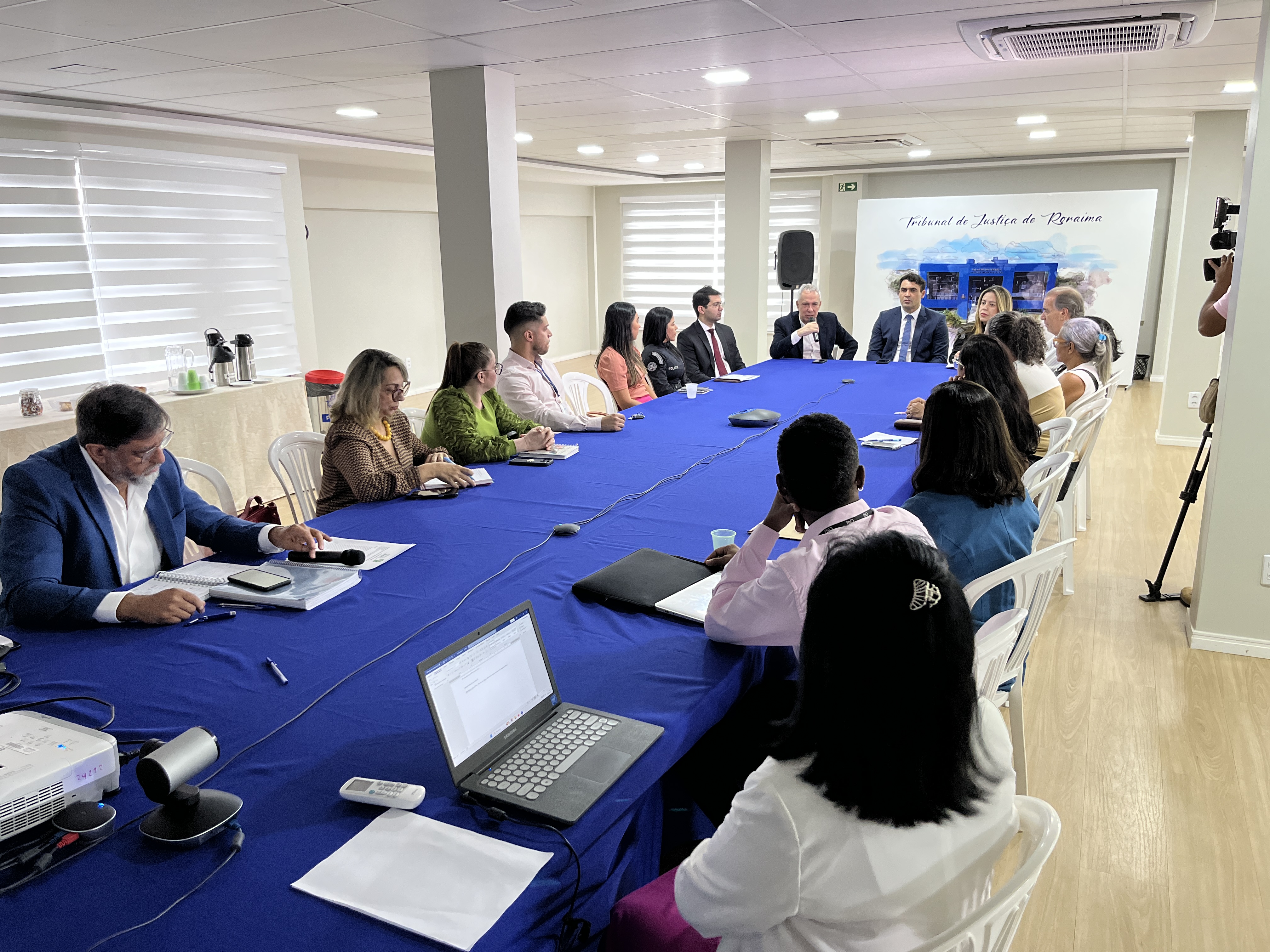 Imagem colorida mostra em ângulo aberto de uma sala de reunião com muitas pessoas sentadas em volta de uma longa mesa retangular com toalha azul. Homens e mulheres participam do encontro, com notebooks e papéis sobre a mesa. Ao fundo, em uma parede branca, há um quadro com a inscrição "Tribunal de Justiça de Roraima". Imagem colorida mostra em ângulo aberto de uma sala de reunião com muitas pessoas sentadas em volta de uma longa mesa retangular com toalha azul. Homens e mulheres participam do encontro, com notebooks e papéis sobre a mesa. Ao fundo, em uma parede branca, há um quadro com a inscrição "Tribunal de Justiça de Roraima".