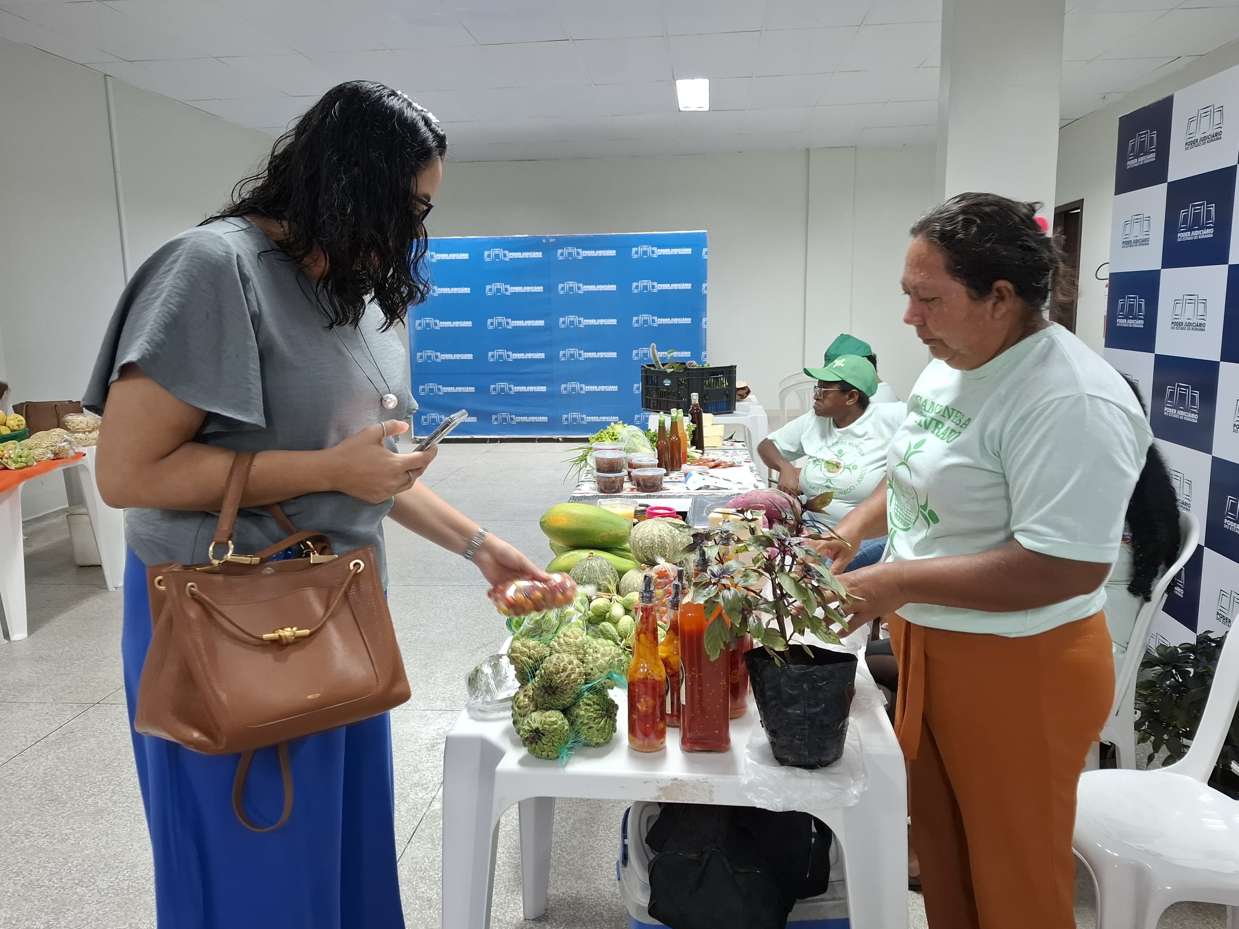 Foto colorida mostra uma consumidora escolhe frutas em uma barraca de feira montada em ambiente interno. Ela observa um pote com tomatinhos enquanto segura o celular e uma bolsa marrom. Do outro lado da mesa, uma expositora de camiseta verde e calça laranja organiza produtos como garrafas de molho, frutas, hortaliças e até mudas de plantas. Ao fundo, um banner azul com o logotipo do Tribunal de Justiça de Roraima. Foto colorida mostra uma consumidora escolhe frutas em uma barraca de feira montada em ambiente interno. Ela observa um pote com tomatinhos enquanto segura o celular e uma bolsa marrom. Do outro lado da mesa, uma expositora de camiseta verde e calça laranja organiza produtos como garrafas de molho, frutas, hortaliças e até mudas de plantas. Ao fundo, um banner azul com o logotipo do Tribunal de Justiça de Roraima.