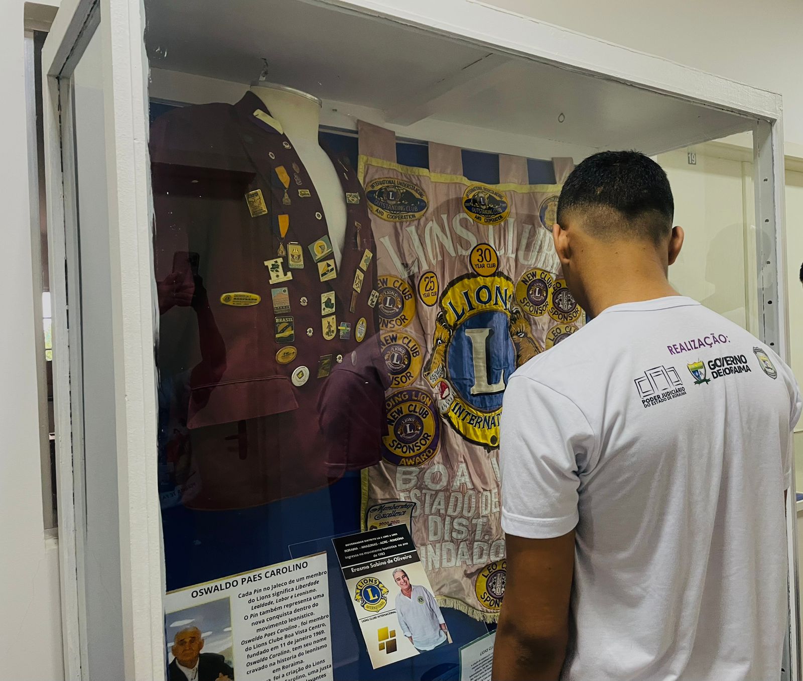 Um homem de costas, vestindo uma camiseta branca, observa um display de vidro. Dentro do display, há um colete de cor escura com vários broches e um estandarte ou bandeira predominantemente azul e dourada com o emblema "LIONS INTERNATIONAL". Há também um texto e uma foto dentro da vitrine, que parece ser um memorial ou exposição. Na parede ao lado do display, há um cartaz com a palavra "REALIZAÇÃO:" e logotipos. Um homem de costas, vestindo uma camiseta branca, observa um display de vidro. Dentro do display, há um colete de cor escura com vários broches e um estandarte ou bandeira predominantemente azul e dourada com o emblema "LIONS INTERNATIONAL". Há também um texto e uma foto dentro da vitrine, que parece ser um memorial ou exposição. Na parede ao lado do display, há um cartaz com a palavra "REALIZAÇÃO:" e logotipos.