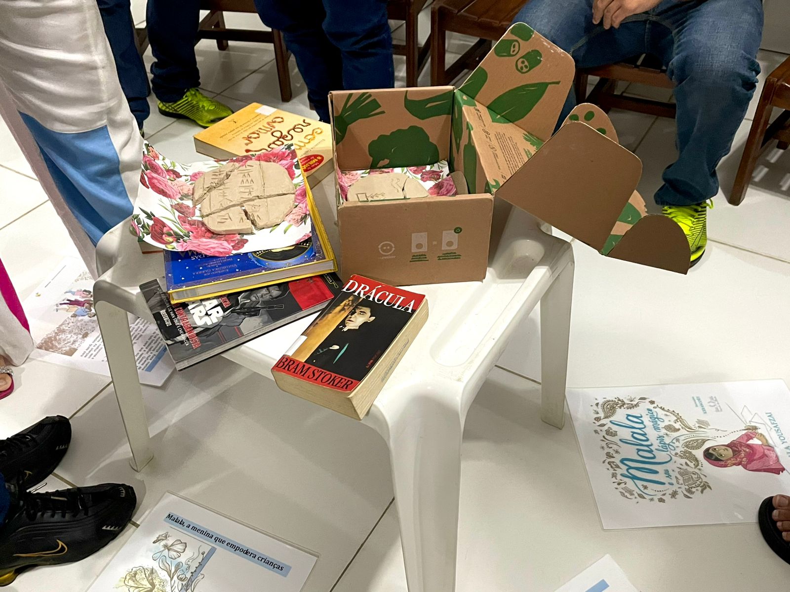 A imagem mostra vários livros dispostos sobre e ao redor de uma pequena mesa branca. Os livros incluem "Drácula" de Bram Stoker e um livro com o título "Malala". Uma caixa de papelão aberta e decorada com folhas verdes também está visível na mesa. Pessoas estão sentadas ao redor da mesa, com os pés e parte das pernas visíveis. O ambiente parece ser interno, possivelmente uma sala. A imagem mostra vários livros dispostos sobre e ao redor de uma pequena mesa branca. Os livros incluem "Drácula" de Bram Stoker e um livro com o título "Malala". Uma caixa de papelão aberta e decorada com folhas verdes também está visível na mesa. Pessoas estão sentadas ao redor da mesa, com os pés e parte das pernas visíveis. O ambiente parece ser interno, possivelmente uma sala.