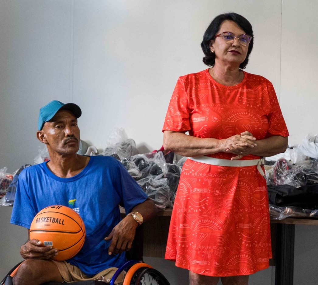 Foto colorida mostra um homem cadeirante com boné azul segurando uma bola de basquete, ao lado da desembargadora Tânia Vasconcelos, que está usando vestido vermelho. Ao fundo, há uma mesa com diversos pares de tênis embalados, sugerindo um evento de doação ou apoio esportivo.