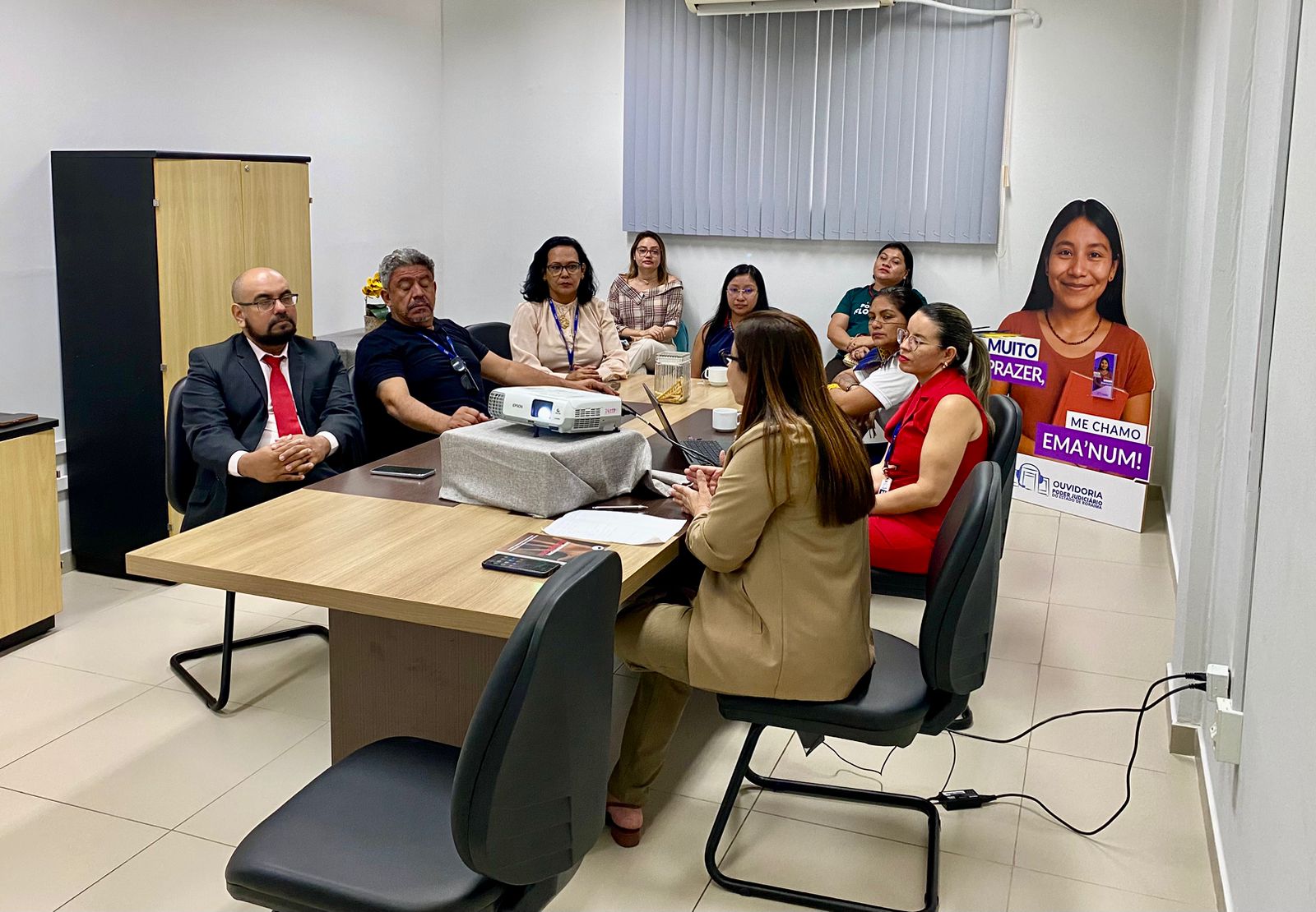 Foto colorida mostra uma reunião em andamento com dez pessoas sentadas em torno de uma mesa de reunião em uma sala do TJRR. No centro da mesa, há um projetor. A maioria dos participantes observa atentamente a mulher que está conduzindo a apresentação. Ao fundo, um totem de papelão em tamanho real exibe a personagem "Ema’num", com a frase: "Muito prazer, me chamo Ema’num!", associada à Ouvidoria do TJRR. 