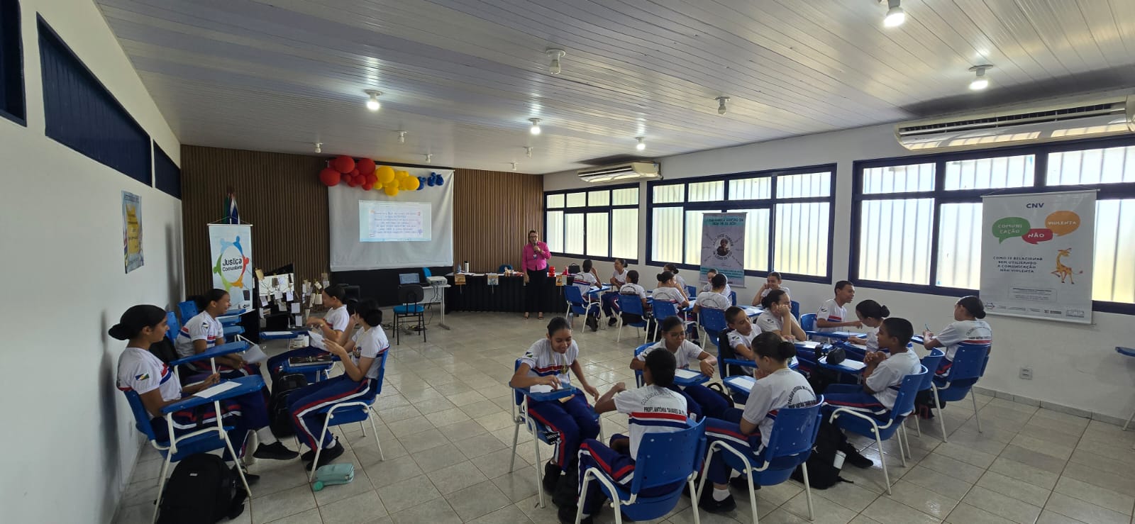 Imagem colorida mostra um grupo de jovens em idade escolar sentados em carteiras de sala de aula, dispostas em grupos de 4, voltados uns para os outros. As carteiras são azuis e brancas. Os jovens estão vestidos com uniformes escolares. Ao fundo, uma mulher adulta está em pé, aparentemente apresentando algo em um projetor que exibe slides em uma tela. Há balões coloridos (vermelho, amarelo e azul) na parede atrás da tela. Na parede direita, há um banner vertical com a inscrição "CNV" (Comunicação Não-Violenta) e palavras como "ouvir", "esperar" e "ação", com um desenho de uma girafa. A sala tem teto claro, luminárias e janelas amplas. Imagem colorida mostra um grupo de jovens em idade escolar sentados em carteiras de sala de aula, dispostas em grupos de 4, voltados uns para os outros. As carteiras são azuis e brancas. Os jovens estão vestidos com uniformes escolares. Ao fundo, uma mulher adulta está em pé, aparentemente apresentando algo em um projetor que exibe slides em uma tela. Há balões coloridos (vermelho, amarelo e azul) na parede atrás da tela. Na parede direita, há um banner vertical com a inscrição "CNV" (Comunicação Não-Violenta) e palavras como "ouvir", "esperar" e "ação", com um desenho de uma girafa. A sala tem teto claro, luminárias e janelas amplas.