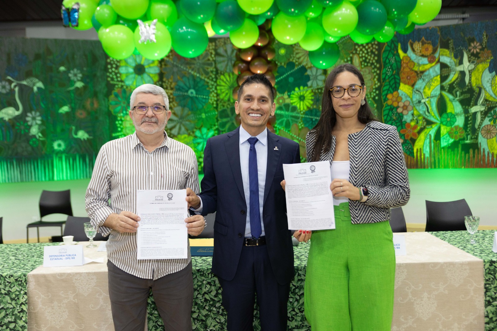Imagem colorida mostra rês pessoas, dois homens e uma mulher, estão sorrindo e segurando documentos. Eles estão de pé atrás de uma mesa coberta por uma toalha verde com padrão de folhas. Ao meio mostra em destaque o juiz coordenador da Coordenadoria da Infância e Juventude e titular da 2ª Vara da Infância e Juventude, Marcelo Oliveira, que usa terno e gravata azul.