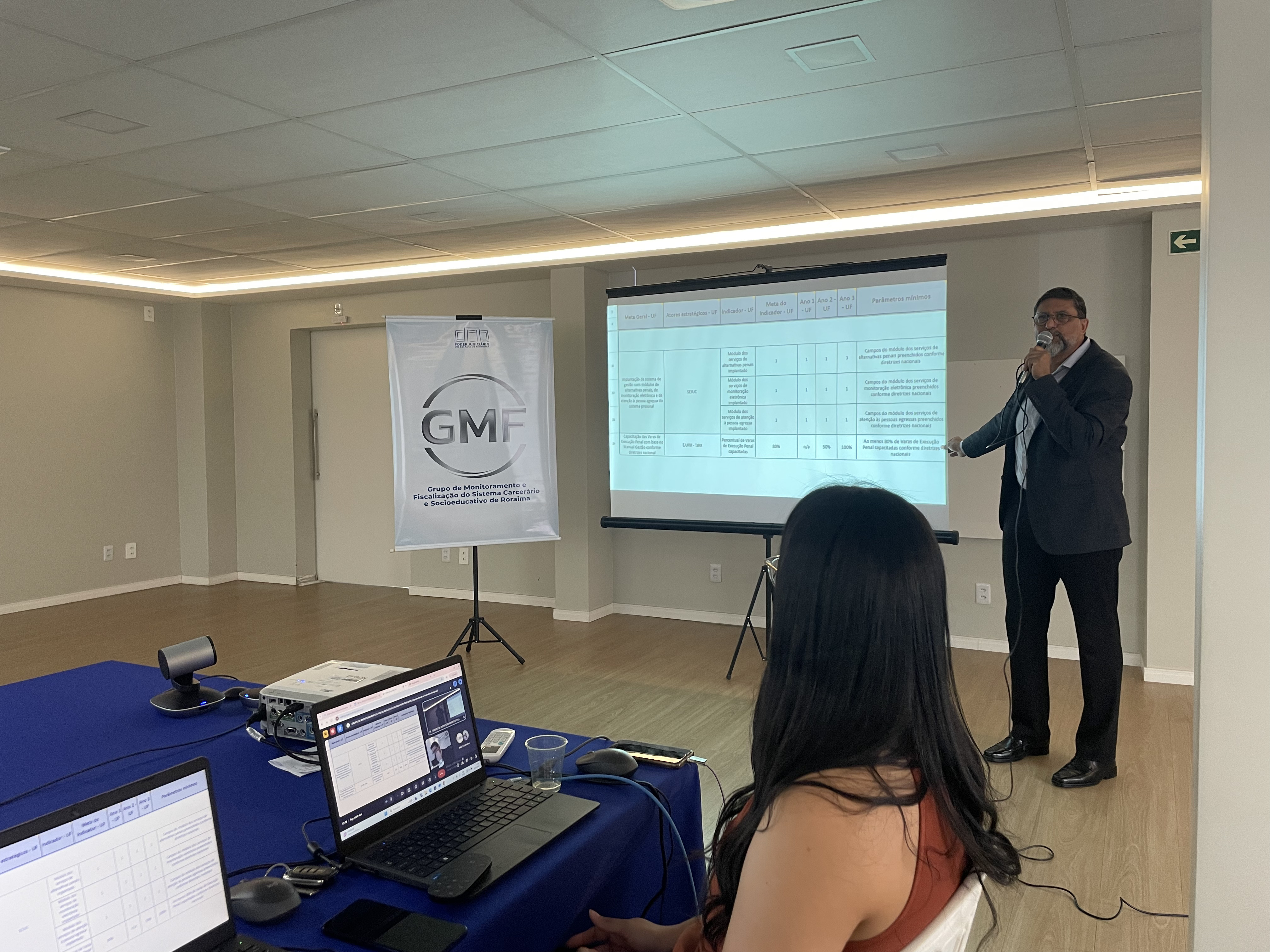 Imagem colorida mostra uma sala de conferências com um homem de terno, Dagoberto Gonçalves, diretor de apoio direto do GMF, em pé ao lado de uma tela de projeção, apontando para ela e falando ao microfone. A tela exibe uma tabela. No lado esquerdo da imagem, há um estande com o logotipo "GMF" e o texto "Grupo de Monitoramento e Fiscalização do Sistema Carcerário e Socioeducativo de Roraima". Uma mulher está sentada em primeiro plano, de costas para a câmera, olhando para a tela. Há também laptops e outros equipamentos eletrônicos sobre uma mesa coberta com um pano azul.