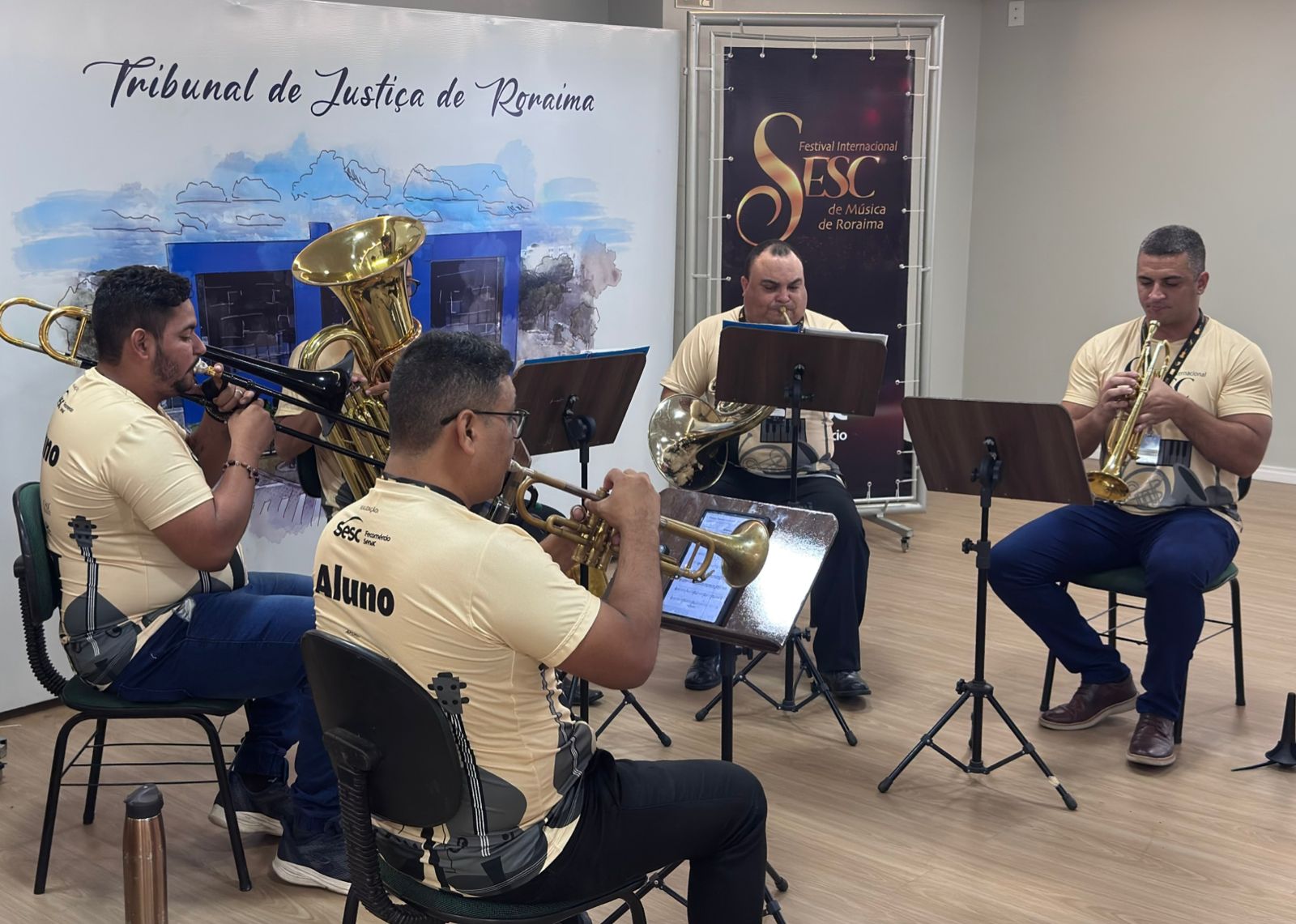 Imagem colorida mostra quatro homens adultos, vestindo camisetas bege do SESC com a estampa de um violino, estão sentados em cadeiras em um ambiente interno. Eles estão tocando instrumentos de sopro de metal: um trombone, uma tuba, uma trombeta e um trompete. Há estantes de partitura em frente a eles. Ao fundo, uma parede branca com a inscrição "Tribunal de Justiça de Roraima" e um banner vertical preto com o logotipo do "Festival Internacional SESC de Música de Roraima".