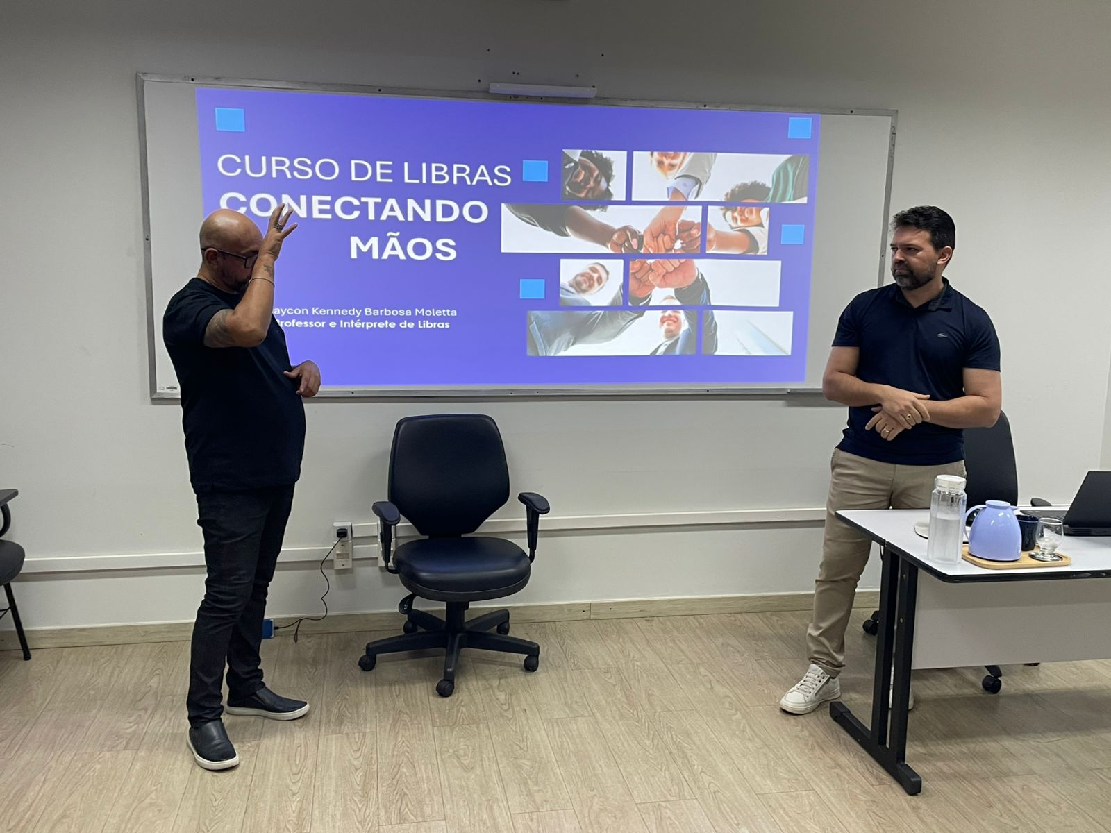 Imagem colorida do professor do curso ao lado de outra pessoa que está fazendo Libras. Imagem colorida do professor do curso ao lado de outra pessoa que está fazendo Libras.