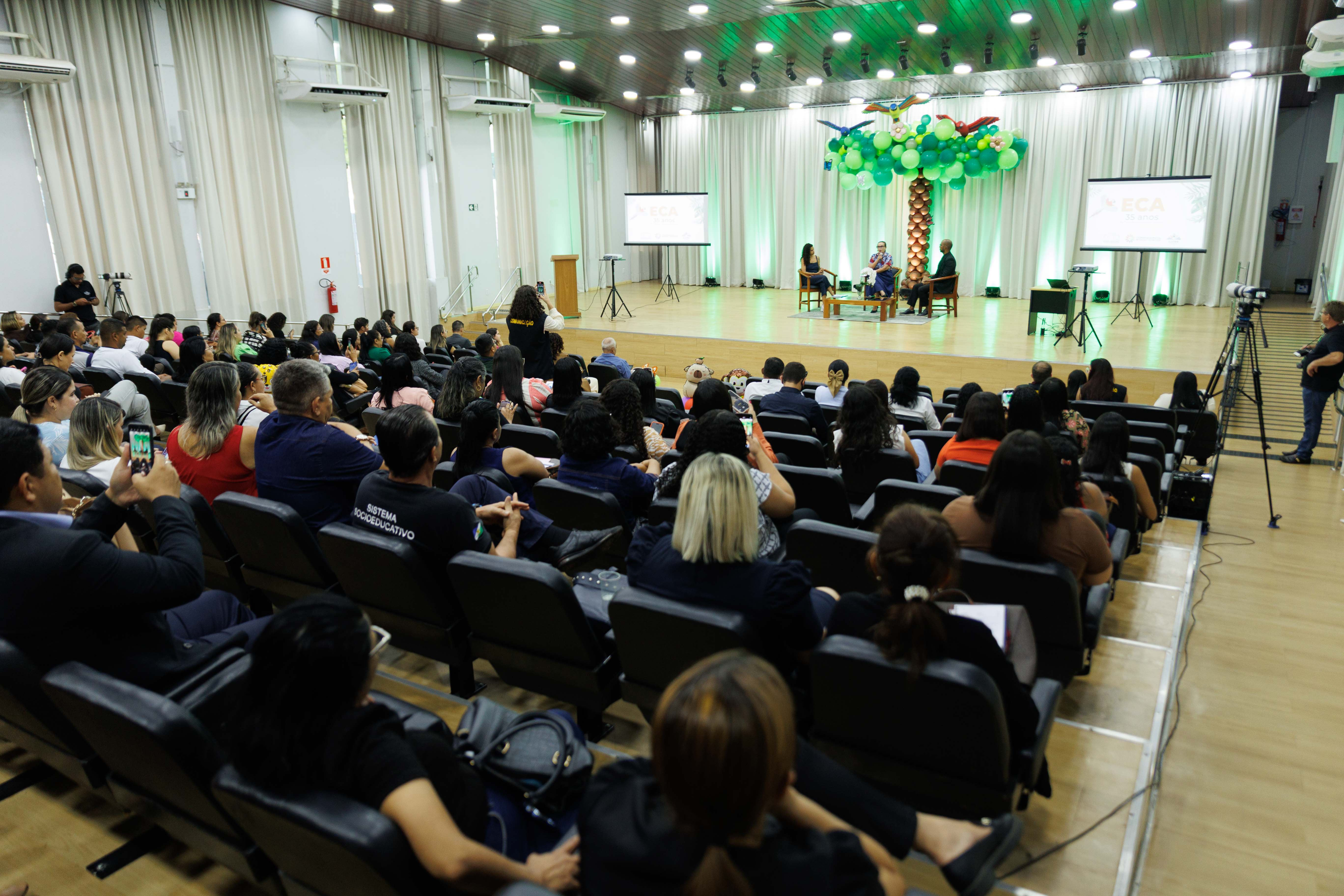 Foto panorâmica de uma roda de conversa durante o evento dos 35 anos do ECA. O auditório está lotado, com dezenas de participantes assistindo atentamente à discussão no palco. Quatro pessoas — três mulheres e um homem — estão sentadas em cadeiras formando um círculo no centro do palco, sob uma grande árvore decorativa feita com balões verdes, flores e araras infláveis. Duas telas laterais projetam a identidade visual do evento.
