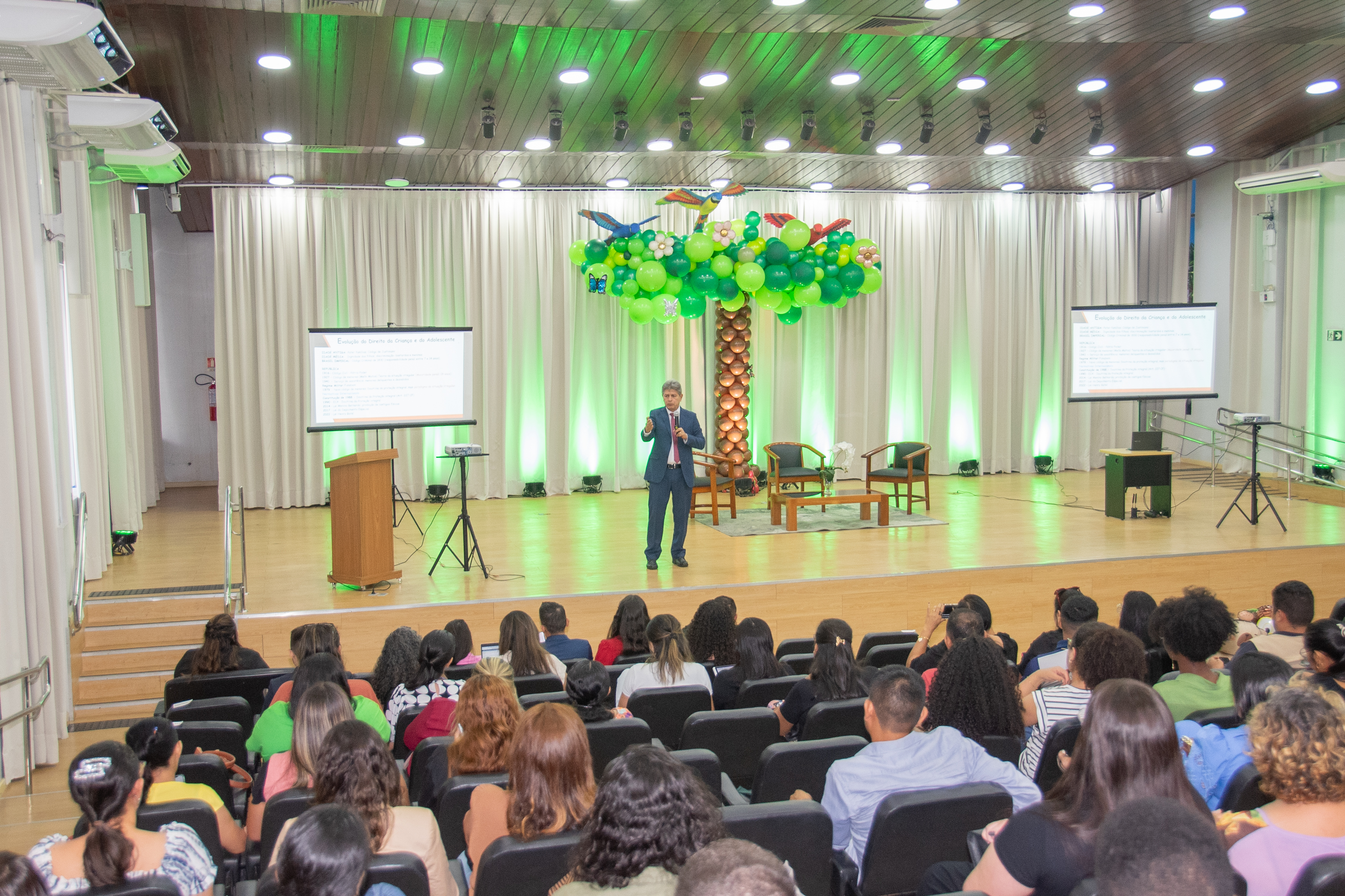 Foto colorida de uma palestra durante o evento comemorativo dos 35 anos do ECA. O juiz titular da 1ª Vara da Infância e Juventude, Parima Dias está no centro do palco, falando ao público com microfone em uma das mãos e a outra erguida. Ao fundo, cortinas brancas e iluminação verde dão o tom do cenário, onde também se destaca uma grande árvore decorativa feita de balões verdes e marrons, com flores e araras infláveis no topo. 