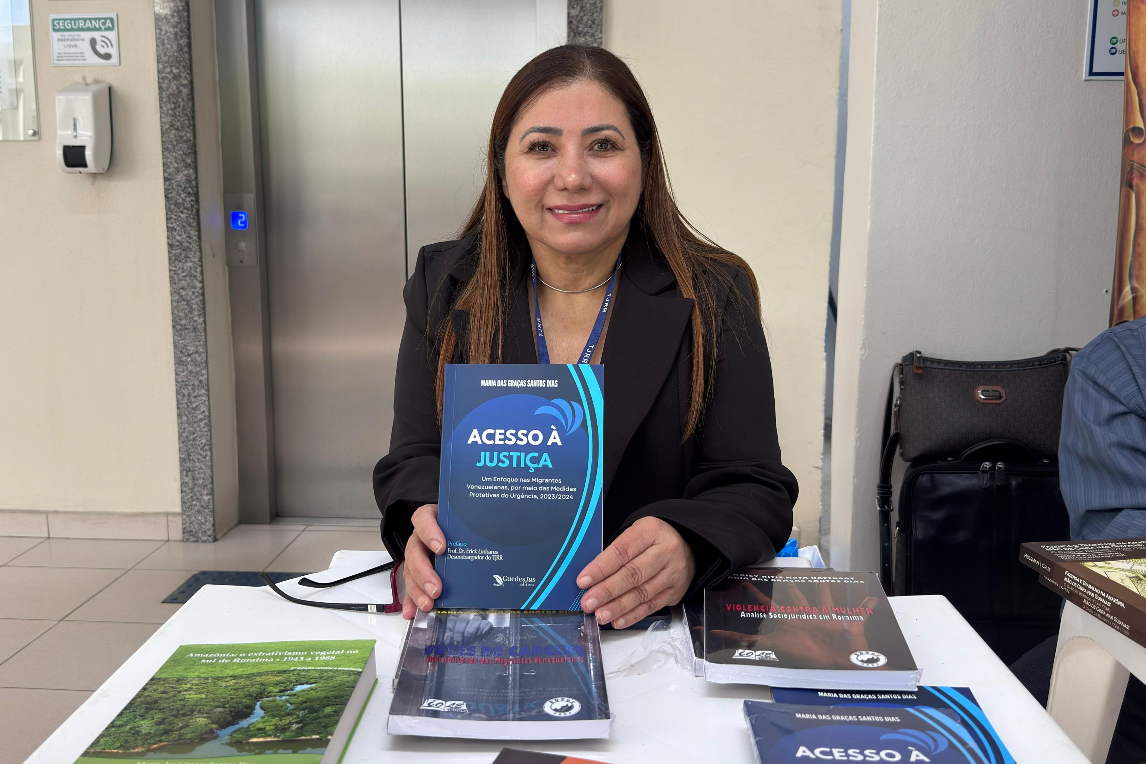Foto colorida mostra uma mulher de cabelos lisos castanhos, trajando blazer preto e crachá institucional do TJRR, sorri enquanto segura o livro "Acesso à Justiça", de sua autoria — Maria das Graças Santos Dias. À sua frente, sobre a mesa, estão outros títulos também voltados à temática dos direitos humanos e justiça. Ao fundo, há um elevador de portas metálicas e paredes claras. A imagem foi registrada durante a Feira do Livro realizada no Tribunal de Justiça de Roraima. Foto colorida mostra uma mulher de cabelos lisos castanhos, trajando blazer preto e crachá institucional do TJRR, sorri enquanto segura o livro "Acesso à Justiça", de sua autoria — Maria das Graças Santos Dias. À sua frente, sobre a mesa, estão outros títulos também voltados à temática dos direitos humanos e justiça. Ao fundo, há um elevador de portas metálicas e paredes claras. A imagem foi registrada durante a Feira do Livro realizada no Tribunal de Justiça de Roraima.