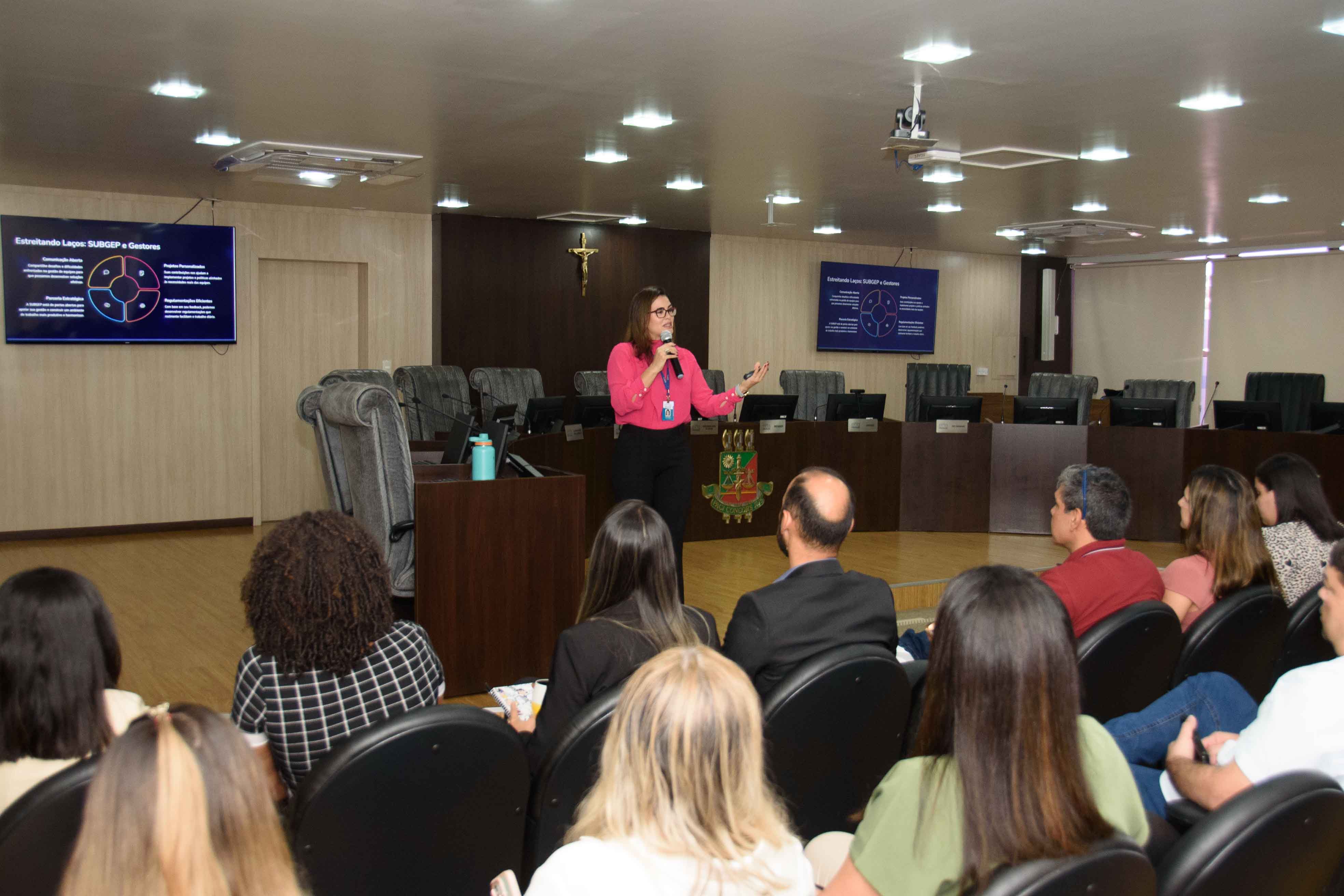 Imagem colorida mostra uma mulher morena de óculos, vestindo uma blusa rosa e calça preta, em pé ao lado de um pódio de madeira, falando e gesticulando com a mão direita. Ao fundo, duas telas exibem slides de apresentação com texto e diagramas, e uma plateia atenta preenche uma sala formal com cadeiras escuras e iluminação de teto.