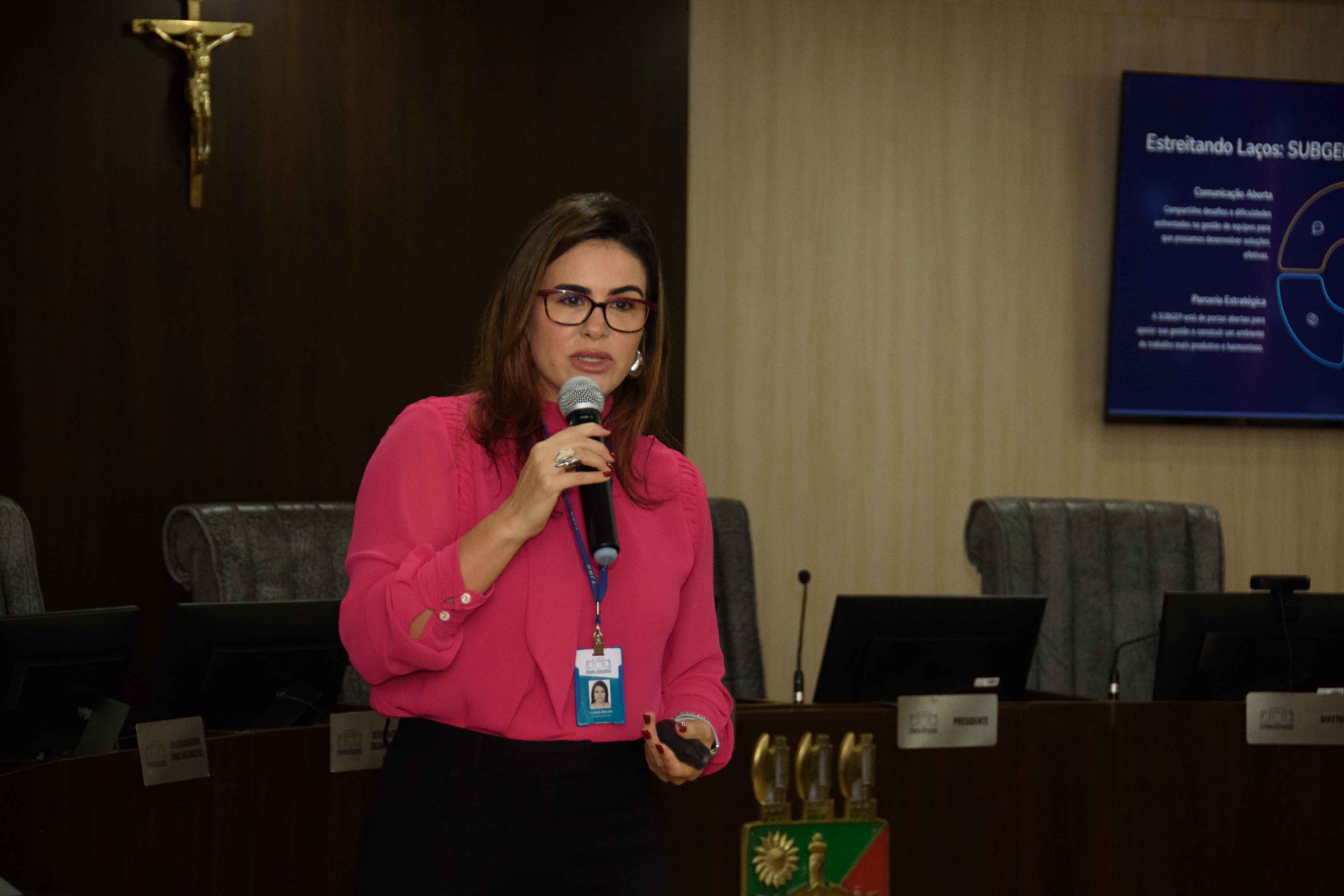Imagem colorida mostra uma mulher morena de óculos, vestindo uma blusa rosa e calça preta, falando em um microfone em um pódio de madeira em frente a uma plateia atenta. Ao fundo, uma tela exibe slides de apresentação com texto e diagramas, em uma sala formal com cadeiras escuras e iluminação de teto.