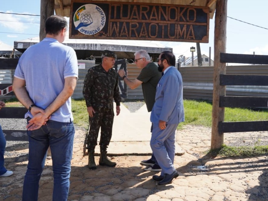  imagem colorida mostra o ministro, o presidente do TJRR, o desembargador Cristovão Súter e um militar em uma área externa, ao ar livre. Ao fundo, há um portão de madeira com uma placa grande e marrom, com o texto "ACOLHIDA ITURANOKO IWARAOTUMA" e "OPERAÇÃO ACOLHIDA".