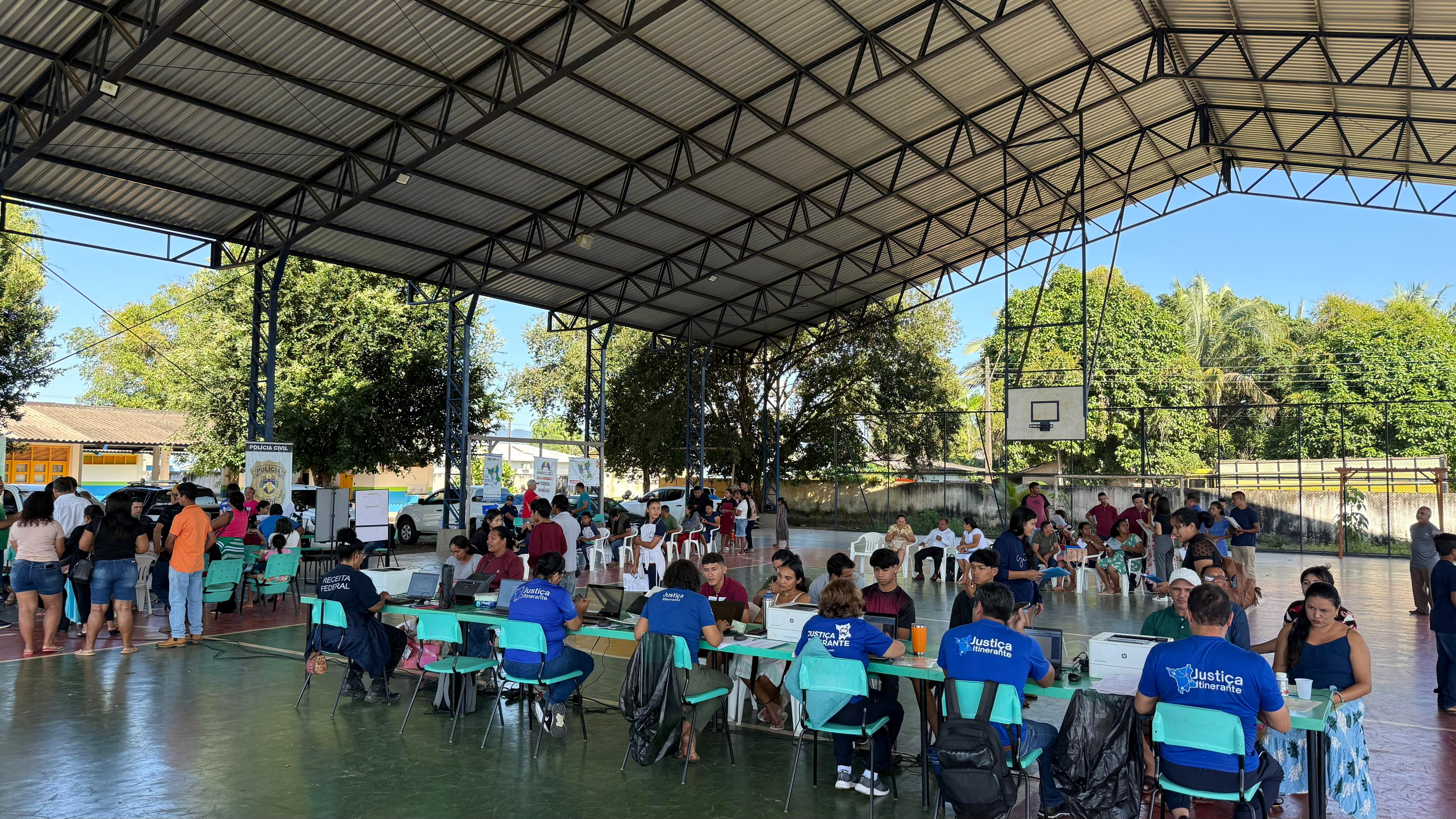Foto colorida mostra um evento de atendimento público social sendo realizado em uma quadra poliesportiva coberta. A quadra tem o chão de cimento pintado de verde. Diversas mesas estão dispostas em filas, com atendentes usando camisetas azuis com a logo "Justiça Itinerante" visível. O público, composto por adultos e crianças, aguarda em cadeiras ou em pé nas filas. Ao fundo, é possível ver árvores e o muro de uma escola. Foto colorida mostra um evento de atendimento público social sendo realizado em uma quadra poliesportiva coberta. A quadra tem o chão de cimento pintado de verde. Diversas mesas estão dispostas em filas, com atendentes usando camisetas azuis com a logo "Justiça Itinerante" visível. O público, composto por adultos e crianças, aguarda em cadeiras ou em pé nas filas. Ao fundo, é possível ver árvores e o muro de uma escola.