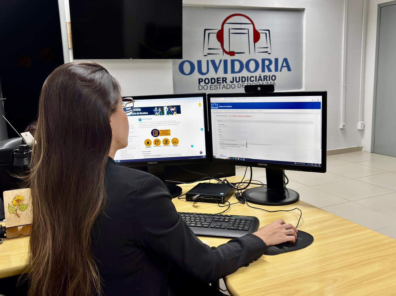 Foto colorida mostra uma mulher trabalhando em dois monitores na Ouvidoria do Poder Judiciário de Roraima. O monitor esquerdo exibe a página principal da ouvidoria, enquanto o direito mostra um formulário online. No fundo, um banner reforça a identidade visual da "OUVIDORIA PODER JUDICIÁRIO DO ESTADO DE RORAIMA".