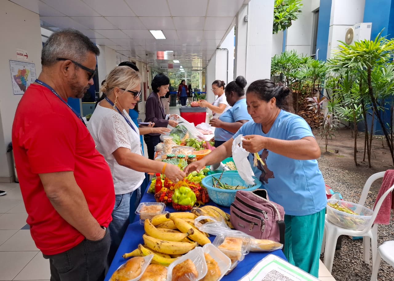 Imagem colorida mostra uma feira com uma mesa de frutas, legumes e verduras, onde clientes estão interagindo com vendedoras. A mesa contém vários cachos de bananas, pimentões, abóboras e outros alimentos em embalagens. O local é um corredor com azulejos brancos e plantas em vasos.