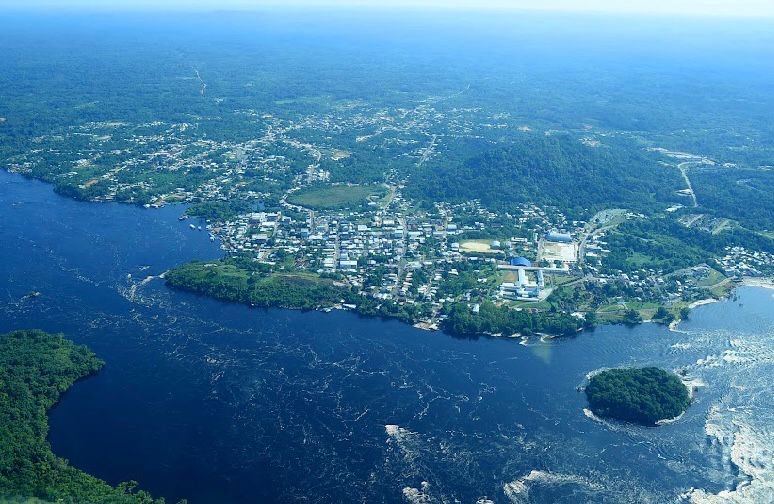 Imagem colorida de vista aérea mostra o município de São Gabriel da Cachoeira (AM) vista de cima. No centro da imagem, observa-se o núcleo urbano com diversas casas, prédios e ruas organizadas. À esquerda, o rio de águas escuras se estende amplamente, com pequenas corredeiras e ilhotas visíveis. À direita, uma região com densa vegetação e morros encobre parte do horizonte Imagem colorida de vista aérea mostra o município de São Gabriel da Cachoeira (AM) vista de cima. No centro da imagem, observa-se o núcleo urbano com diversas casas, prédios e ruas organizadas. À esquerda, o rio de águas escuras se estende amplamente, com pequenas corredeiras e ilhotas visíveis. À direita, uma região com densa vegetação e morros encobre parte do horizonte