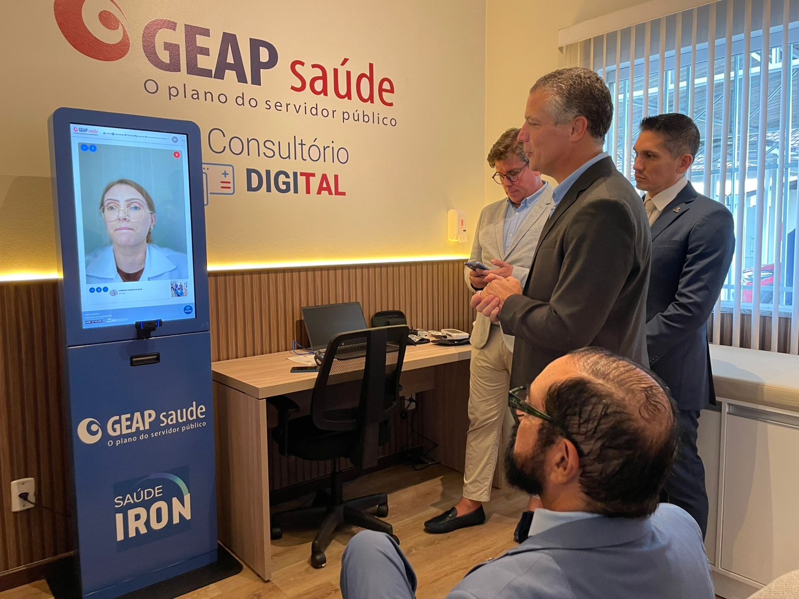  imagem colorida do Desembargador Leonardo Cupello participando de demonstração do atendimento remoto durante a inauguração do consultório digital Geap Saúde no TJRR, a frente há uma médica em um totem.