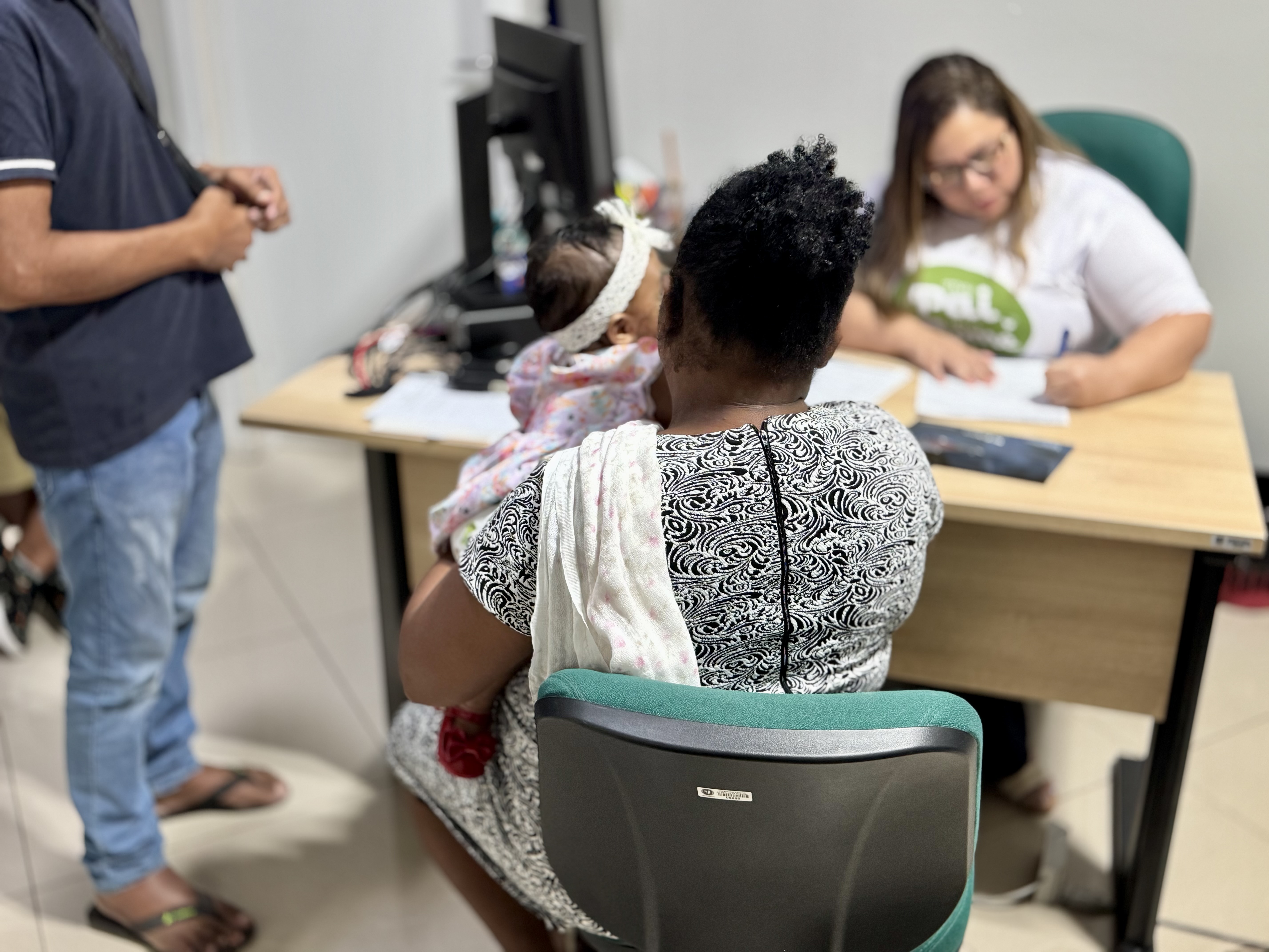 Imagem colorida mostra uma mulher negra de costas, sentada em uma cadeira cinza e verde, segurando um bebê que usa uma faixa de cabeça branca. A mulher veste um vestido preto e branco. À direita, outra mulher, branca e de cabelos castanhos, sentada em uma mesa de escritório, olha para baixo enquanto escreve em uma folha de papel. À esquerda, um homem negro, de costas, veste uma camisa polo azul escura e jeans. O ambiente é um escritório com um computador na mesa. Imagem colorida mostra uma mulher negra de costas, sentada em uma cadeira cinza e verde, segurando um bebê que usa uma faixa de cabeça branca. A mulher veste um vestido preto e branco. À direita, outra mulher, branca e de cabelos castanhos, sentada em uma mesa de escritório, olha para baixo enquanto escreve em uma folha de papel. À esquerda, um homem negro, de costas, veste uma camisa polo azul escura e jeans. O ambiente é um escritório com um computador na mesa.