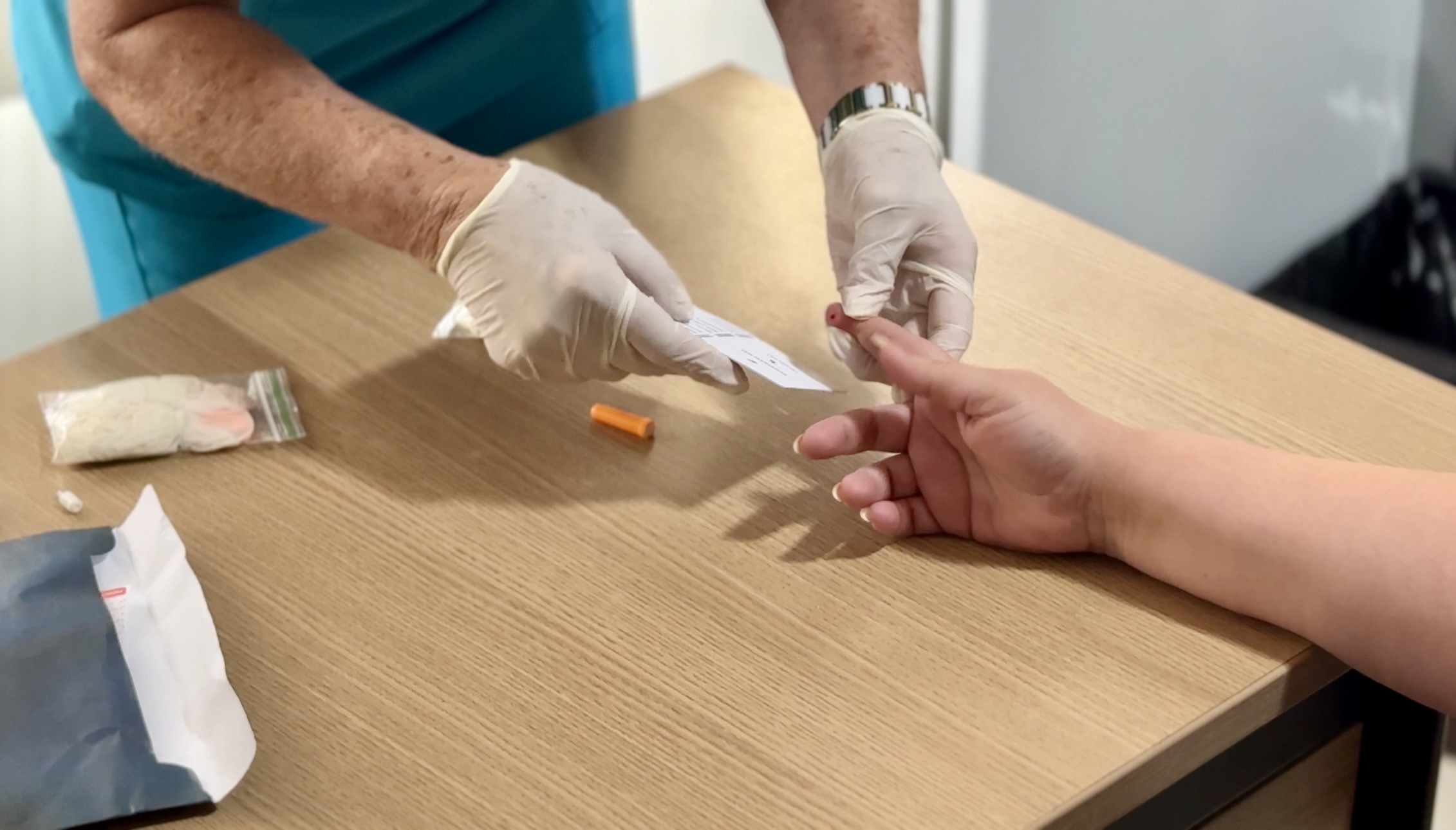 Imagem colorida mostra as mãos de uma pessoa de avental azul e luvas brancas, perfurando o dedo de outra pessoa para coleta de sangue. Um pequeno papel branco com um código de barras está sendo segurado próximo ao dedo. Um kit para teste, incluindo uma bandagem e um pequeno tubo laranja, está sobre a mesa de madeira clara. Imagem colorida mostra as mãos de uma pessoa de avental azul e luvas brancas, perfurando o dedo de outra pessoa para coleta de sangue. Um pequeno papel branco com um código de barras está sendo segurado próximo ao dedo. Um kit para teste, incluindo uma bandagem e um pequeno tubo laranja, está sobre a mesa de madeira clara.
