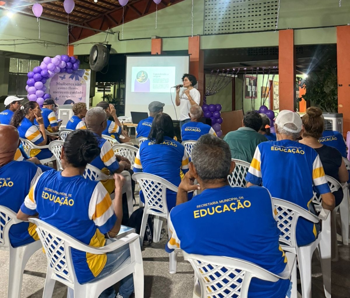 Fotografia mostra uma palestra realizada em um espaço fechado. No centro, uma mulher em pé, usando camiseta branca, fala ao público com microfone em mãos, em frente a uma tela de projeção que exibe um slide. O auditório está decorado com balões lilás e uma faixa onde se lê: “Mulheres são como flores: merecem cuidado e respeito”. As cadeiras brancas estão ocupadas por participantes, a maioria vestindo camisas azuis com detalhes amarelos e brancos, onde está escrito “Secretaria Municipal de Educação”.