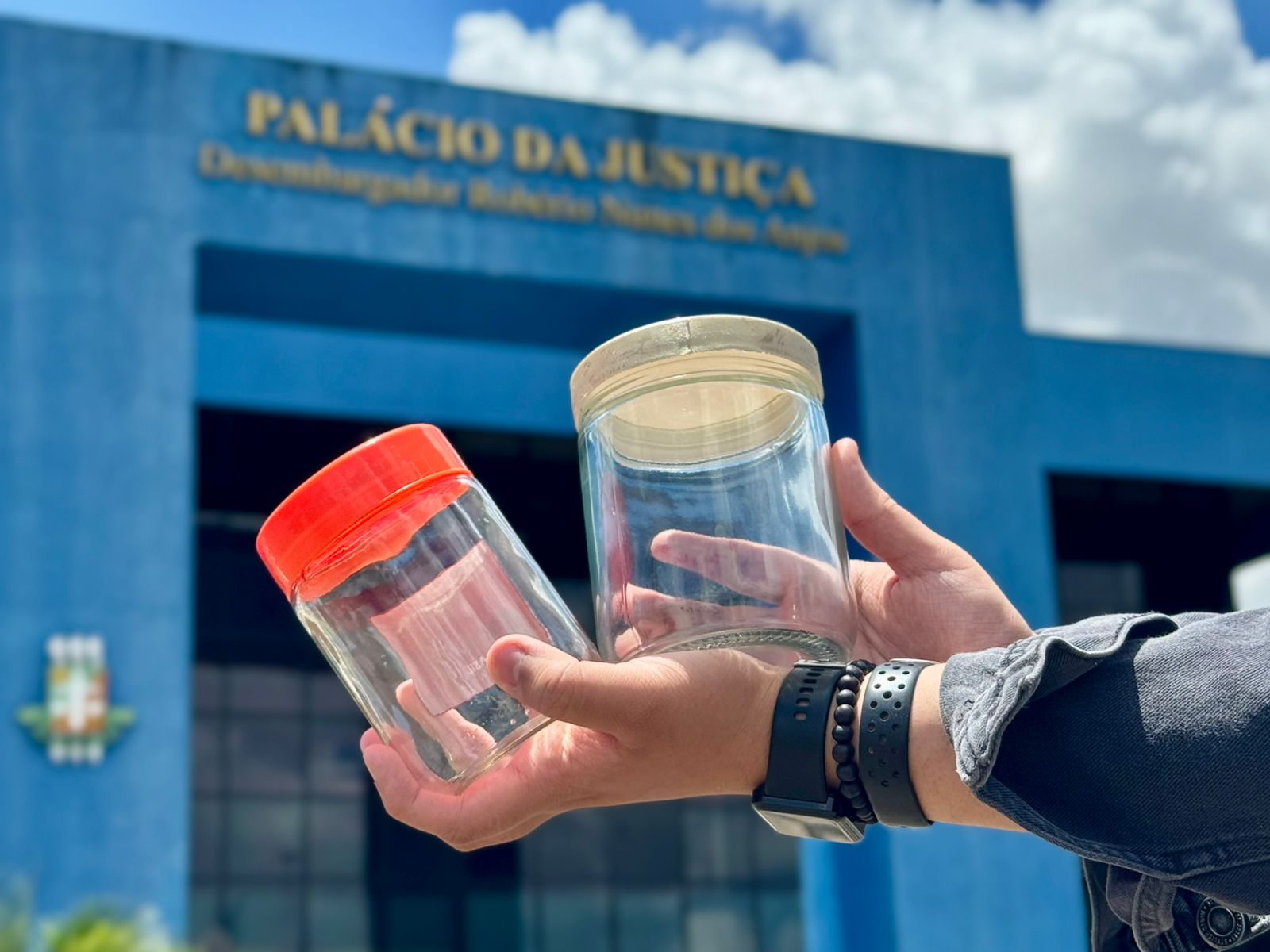 Imagem colorida mostra duas mãos seguram dois potes de vidro, um com tampa laranja e outro com tampa clara, em frente ao Palácio da Justiça Desembargador Robério Nunes dos Anjos do TJRR. Imagem colorida mostra duas mãos seguram dois potes de vidro, um com tampa laranja e outro com tampa clara, em frente ao Palácio da Justiça Desembargador Robério Nunes dos Anjos do TJRR.