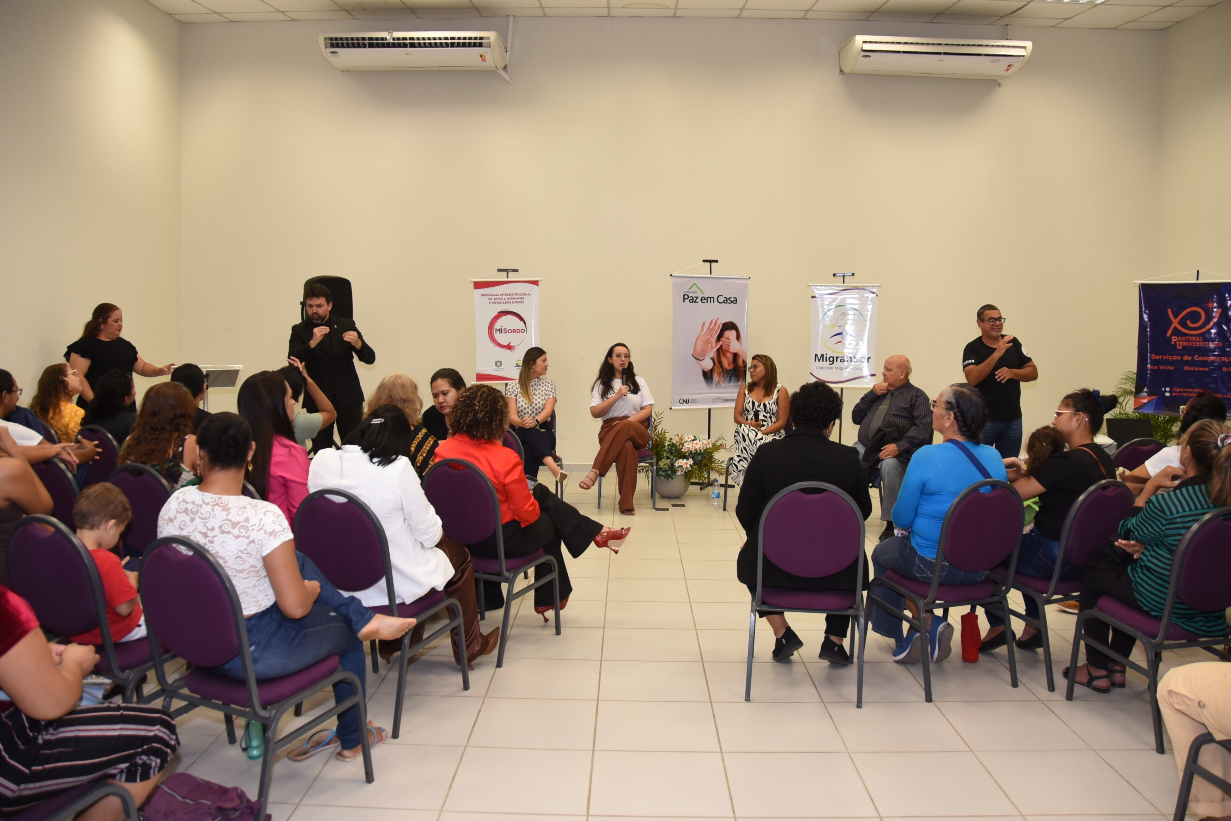 Foto colorida mostra um auditório com cadeiras dispostas em semicírculo, onde um público diverso acompanha uma roda de conversa com quatro pessoas sentadas à frente. Da esquerda para a direita estão, a delegada, Clarissa Pinheiro, a juíza Suelen Alves, a representante do projeto Mujeres: Sinais de Alerta, Thaisy Bentes e o Defensor Público, Wallace Rodrigues. Em pé, à direita está o intereprete de Libras e a esquerda o interprete de LSV. Foto colorida mostra um auditório com cadeiras dispostas em semicírculo, onde um público diverso acompanha uma roda de conversa com quatro pessoas sentadas à frente. Da esquerda para a direita estão, a delegada, Clarissa Pinheiro, a juíza Suelen Alves, a representante do projeto Mujeres: Sinais de Alerta, Thaisy Bentes e o Defensor Público, Wallace Rodrigues. Em pé, à direita está o intereprete de Libras e a esquerda o interprete de LSV.