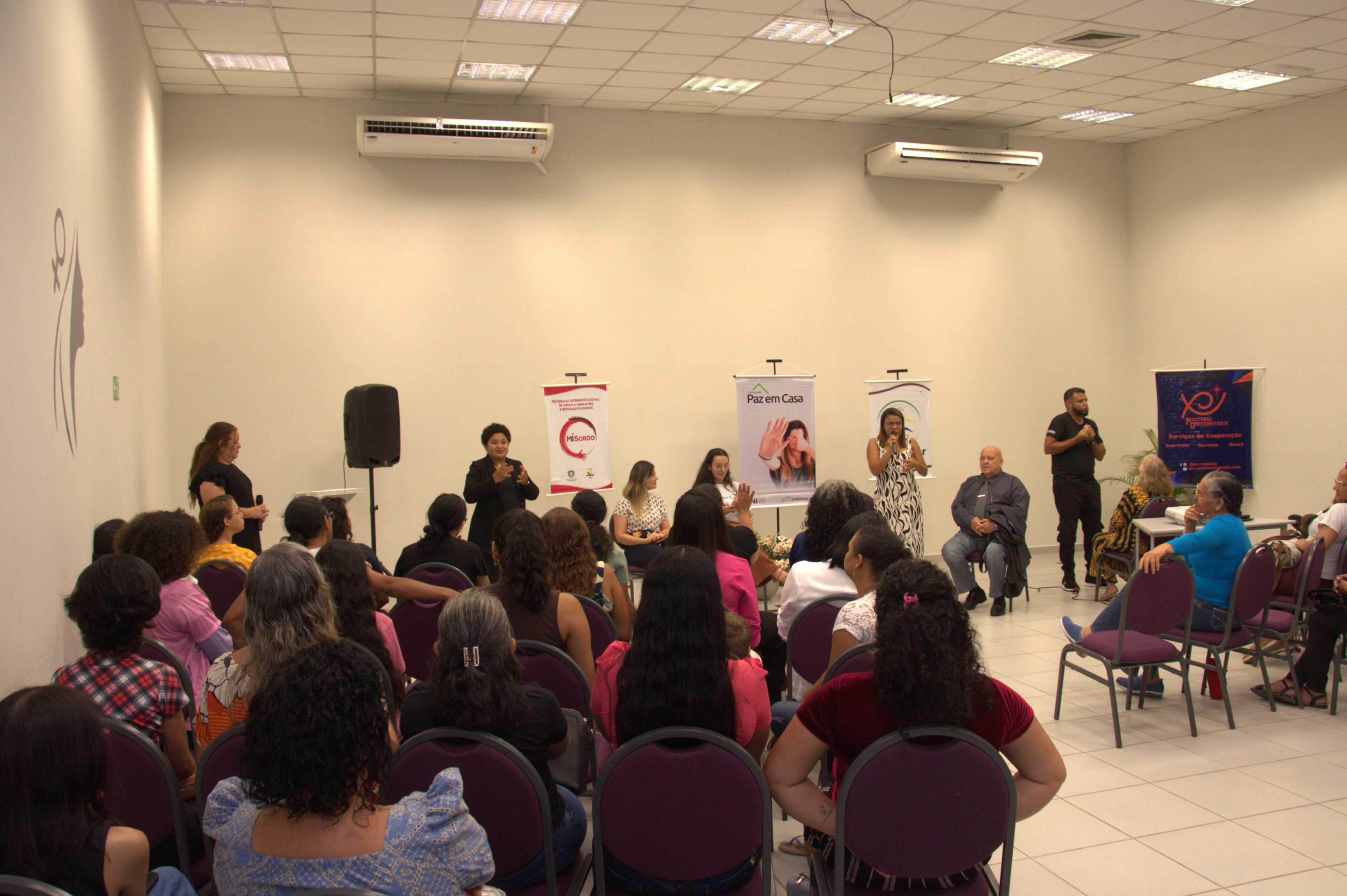 Foto colorida mostra uma palestra realizada em auditório. O público é composto majoritariamente por mulheres, que assistem à apresentação de um grupo posicionado à frente. No palco, há três pessoas sentadas e quatro em pé, sendo duas intérpretes de linguagem de sinal — uma à esquerda, interpretando em Libras e outra à direita da imagem, interpretando em LSV. Foto colorida mostra uma palestra realizada em auditório. O público é composto majoritariamente por mulheres, que assistem à apresentação de um grupo posicionado à frente. No palco, há três pessoas sentadas e quatro em pé, sendo duas intérpretes de linguagem de sinal — uma à esquerda, interpretando em Libras e outra à direita da imagem, interpretando em LSV.