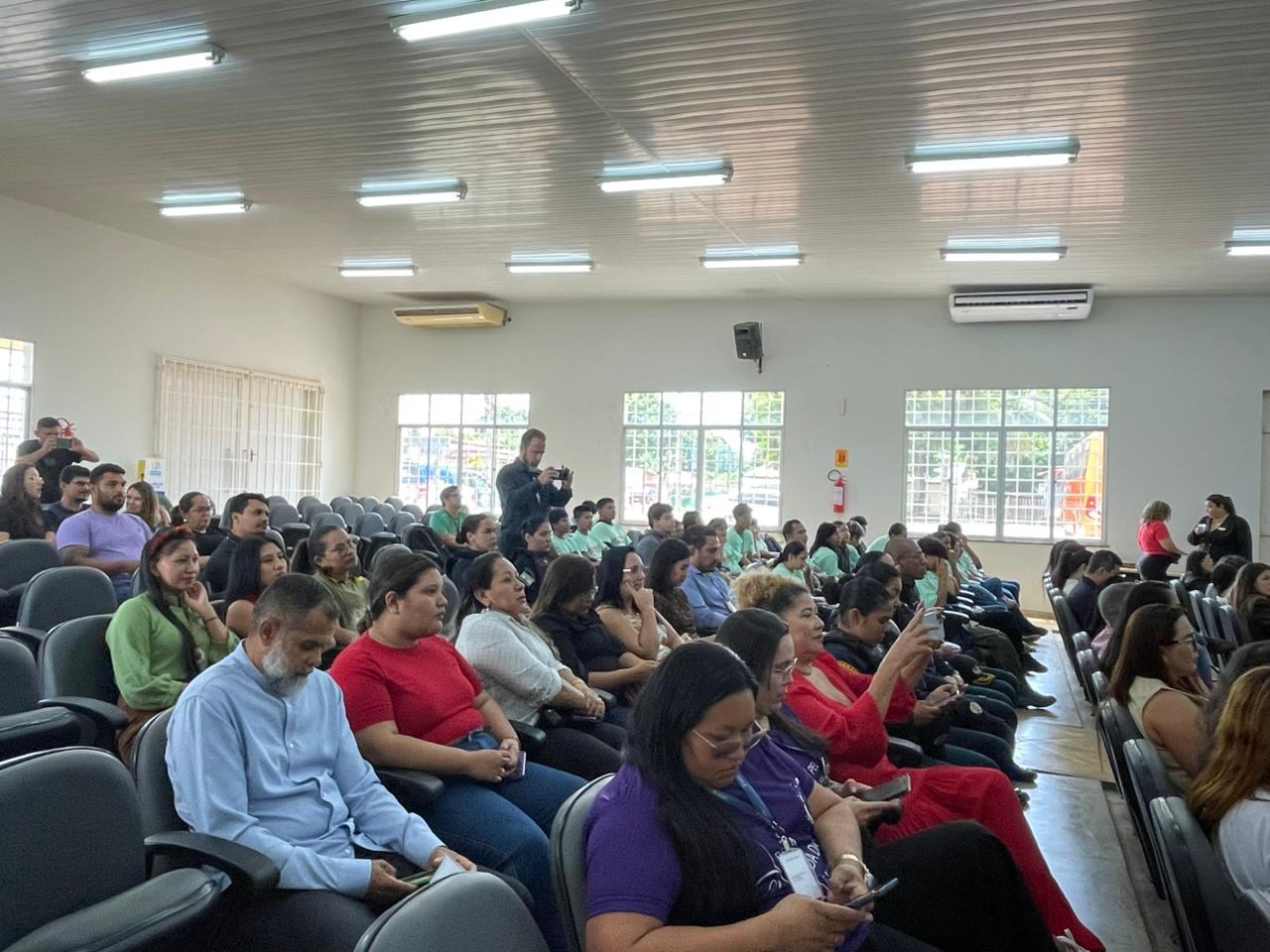 Foto colorida mostra a plateia do evento. As pessoas estão sentadas, prestando atenção às falas, algumas registrando o momento com celulares. O auditório é amplo e tem grandes janelas laterais. Foto colorida mostra a plateia do evento. As pessoas estão sentadas, prestando atenção às falas, algumas registrando o momento com celulares. O auditório é amplo e tem grandes janelas laterais.