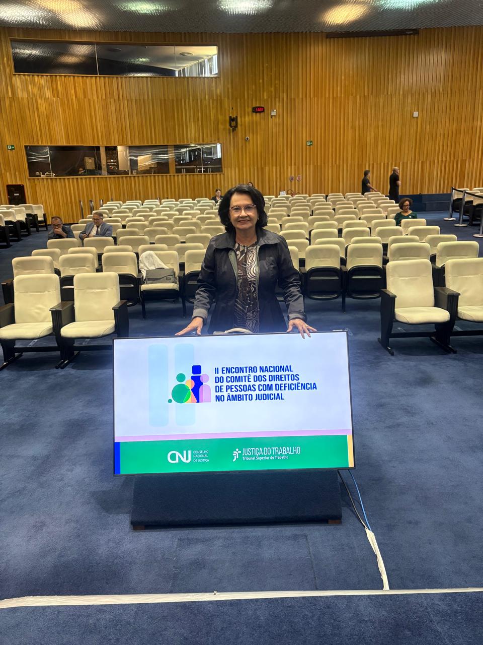 Imagem colorida mostra a desembargadora Tânia Vasconcelos em pé na frente de uma tela digital grande. A tela exibe o título "II ENCONTRO NACIONAL DO COMITÊ DOS DIREITOS DE PESSOAS COM DEFICIÊNCIA NO ÂMBITO JUDICIAL" e os logotipos do CNJ e da Justiça do Trabalho. Ela está em um auditório com fileiras de cadeiras vazias. A parede do fundo é de madeira, e o chão é revestido de carpete azul. Imagem colorida mostra a desembargadora Tânia Vasconcelos em pé na frente de uma tela digital grande. A tela exibe o título "II ENCONTRO NACIONAL DO COMITÊ DOS DIREITOS DE PESSOAS COM DEFICIÊNCIA NO ÂMBITO JUDICIAL" e os logotipos do CNJ e da Justiça do Trabalho. Ela está em um auditório com fileiras de cadeiras vazias. A parede do fundo é de madeira, e o chão é revestido de carpete azul.